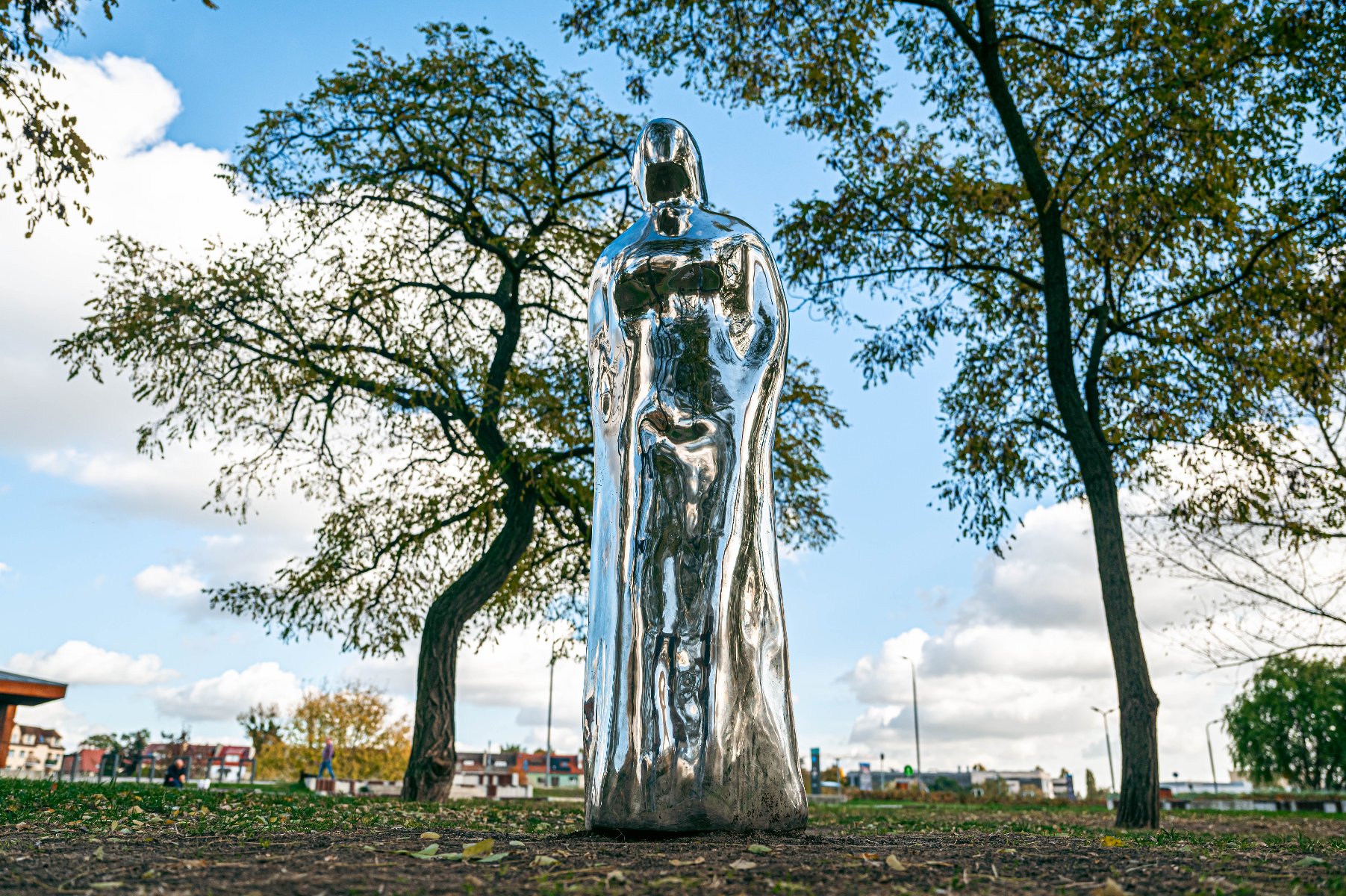 Metaliczna, błyszcząca statua w parku. Przedstawia ludzką postać w płaszczu, o gładkiej, lustrzanej powierzchni, która odbija otaczającą przestrzeń. - grafika artykułu