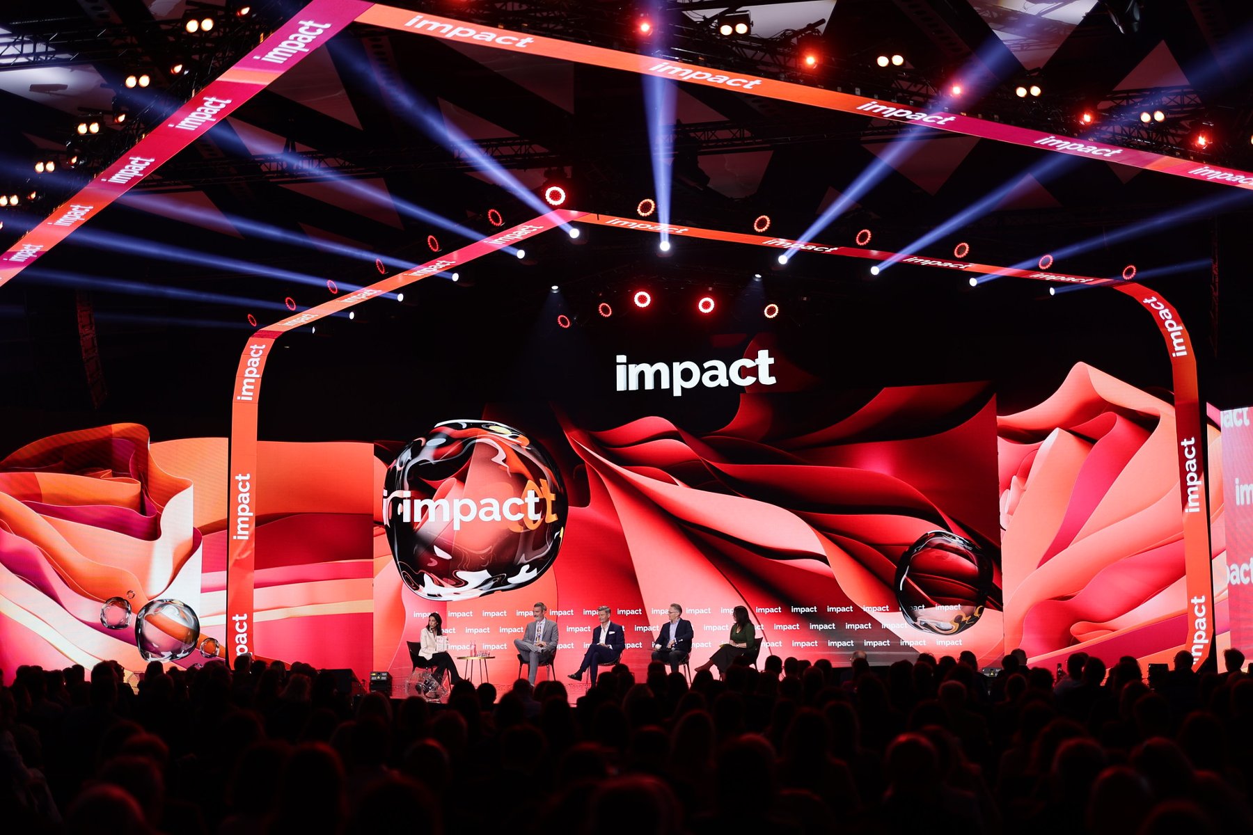 Scena konferencyjna z dużym ekranem wyświetlającym wielokrotnie słowo "impact". Na scenie pięć osób siedzących na krzesłach, prawdopodobnie uczestniczących w panelu dyskusyjnym. Tło w odcieniach czerwieni i pomarańczu, kilka kulistych obiektów także z napisem "impact". Dynamiczne oświetlenie skierowane na scenę, w tle widoczna publiczność - grafika artykułu