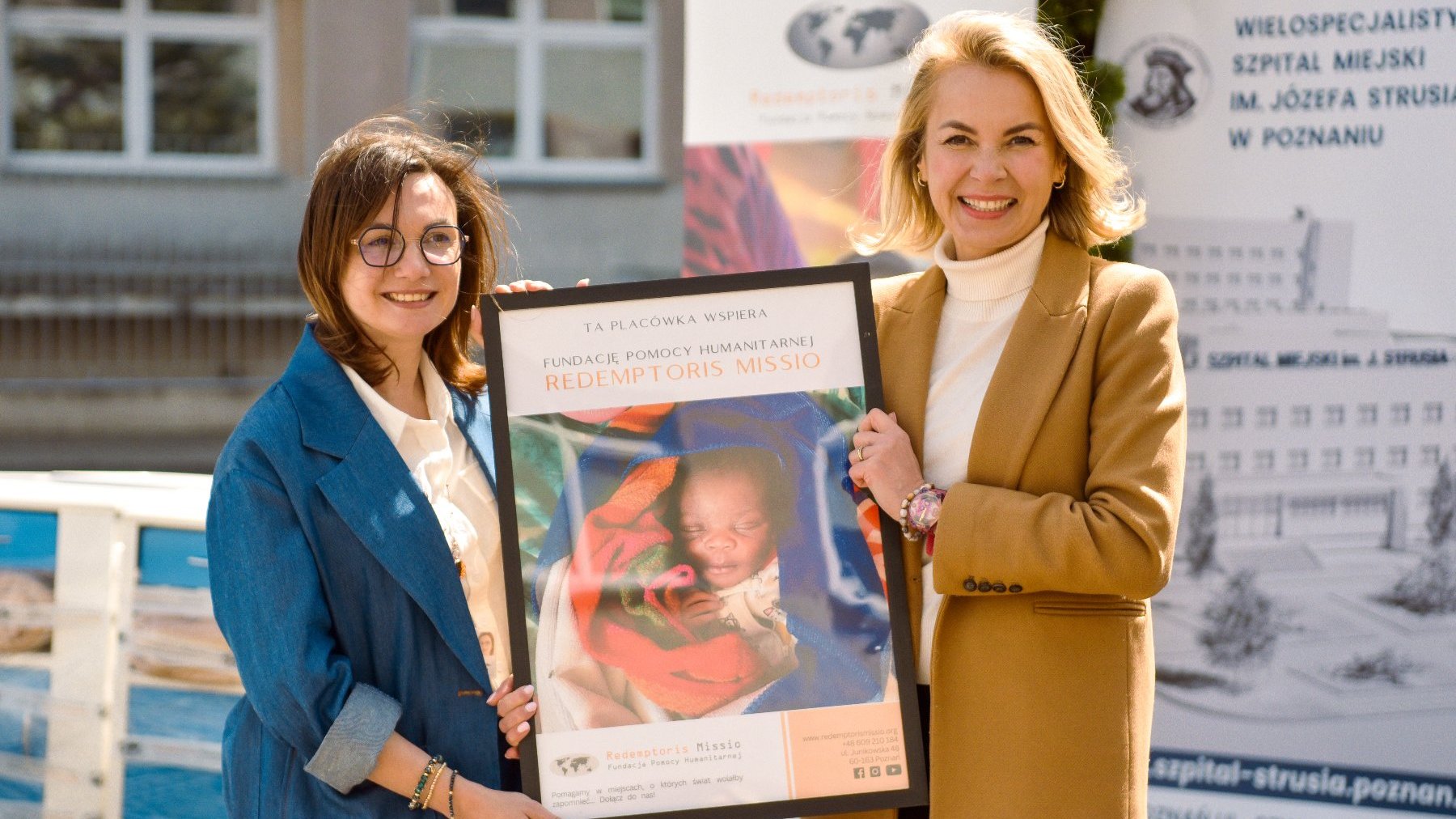 Na zdjęciu dwie kobiety trzymające plakat, na nim małe dziecko