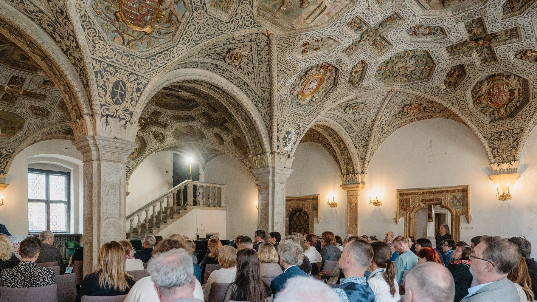 Zdjęcie przedstawia tłum ludzi siedzących wewnątrz ratuszowej sali.