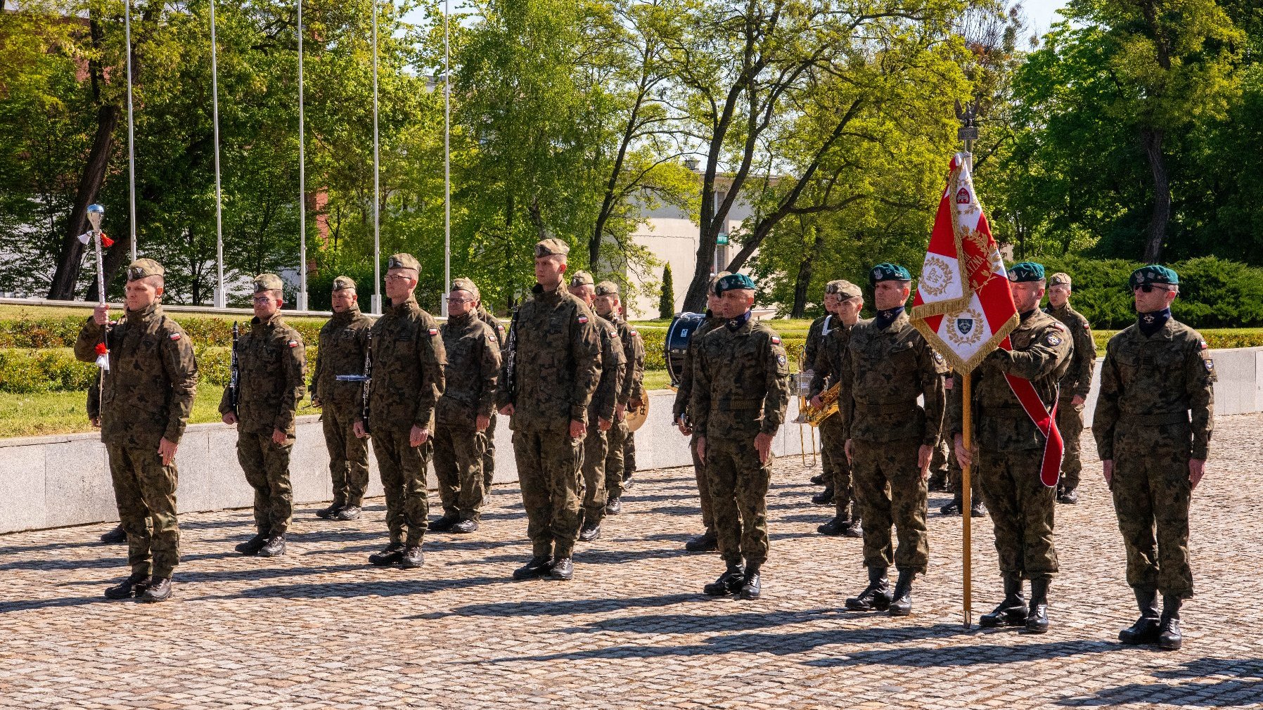 Grupa żołnierzy stoi w szyku na świeżym powietrzu. Mają na sobie mundury maskujące i berety. Jeden z żołnierzy trzyma flagę, a inny ceremonialny miecz. W tle widoczne są maszty z czerwono-białymi flagami. Otoczenie przypomina park, z drzewami i zielenią.
