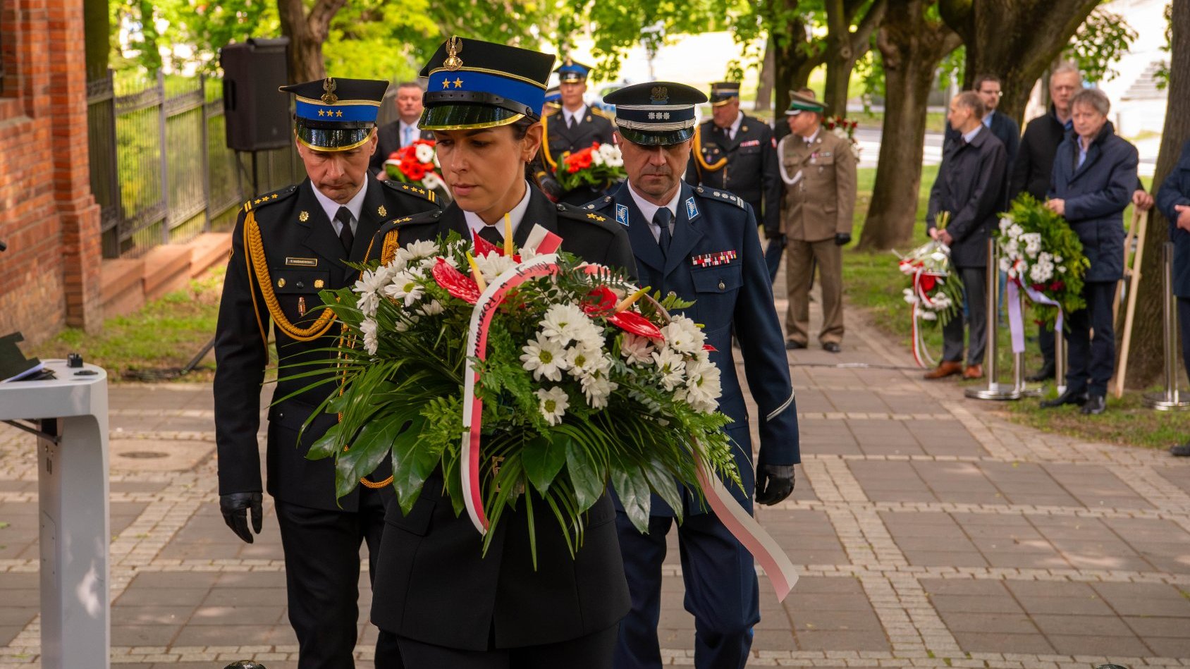 Grupa osób uczestniczących w ceremonii na świeżym powietrzu. Na pierwszym planie osoba w mundurze trzyma duży wieniec z białymi kwiatami, zielonymi liśćmi i czerwonymi akcentami, przewiązany wstążką.
