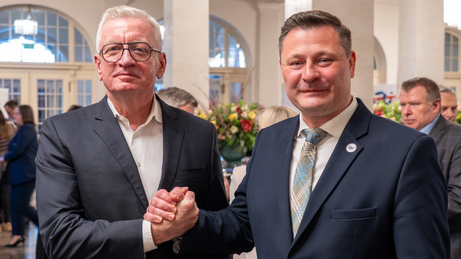Dwóch mężczyzn w eleganckich garniturach podaje sobie rękę. Jeden ma na sobie ciemny garnitur z białą koszulą, drugi dodatkowo nosi wzorzysty krawat. W tle dobrze oświetlone pomieszczenie z dużymi oknami.