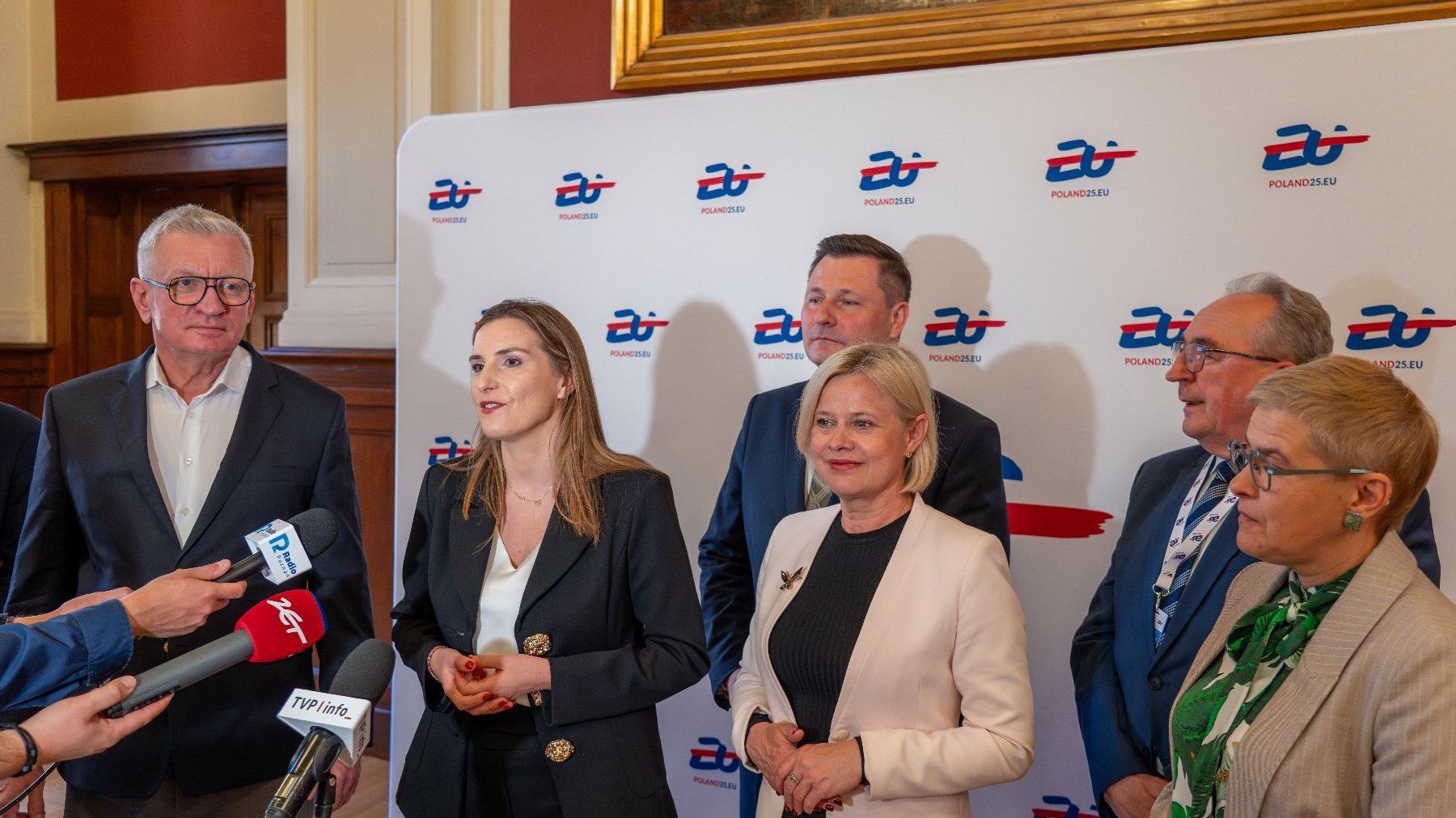 Grupa osób stoi przed białym tłem z powtarzającym się logo "Poland2050.eu'. W tle widać drewniane panele i oprawione obrazy, sugerujące oficjalne wnętrze. Kilka osób trzyma mikrofony.