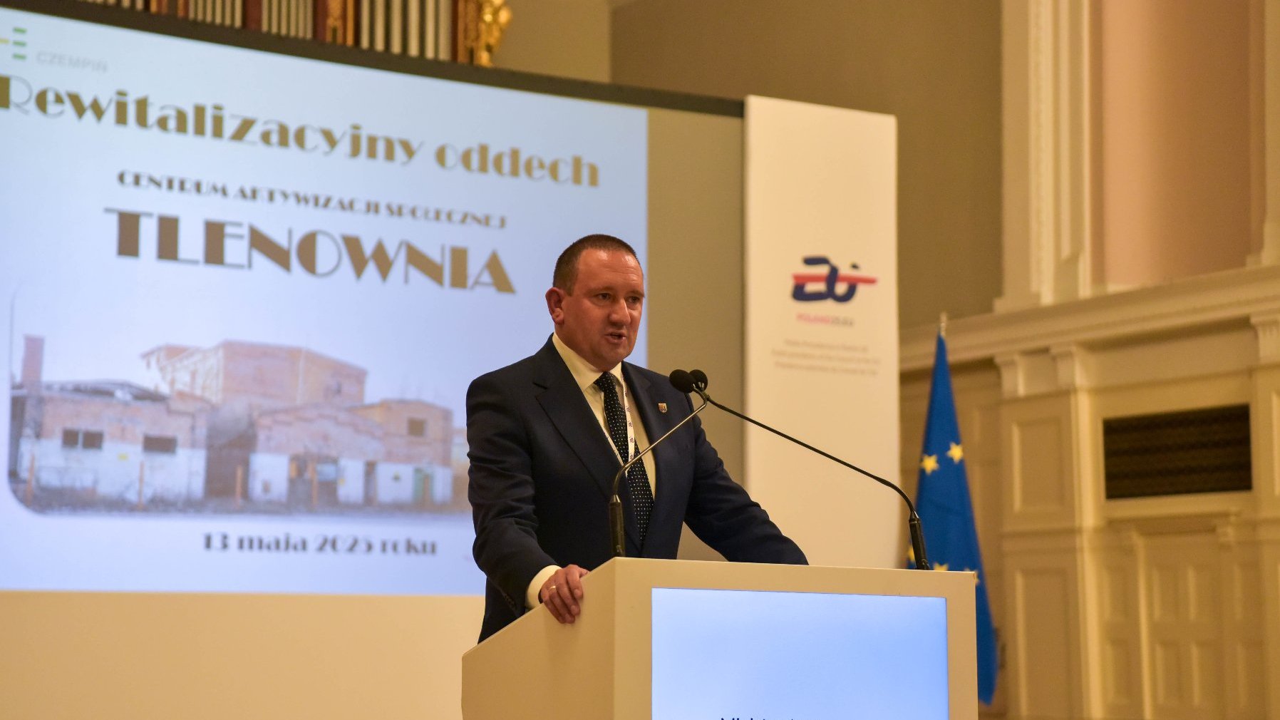 Mężczyzna stoi przy podium z napisem "Ministerstwo Rozwoju i Technologii'. Na dużym ekranie za nim widnieje prezentacja z grafiką budynku.