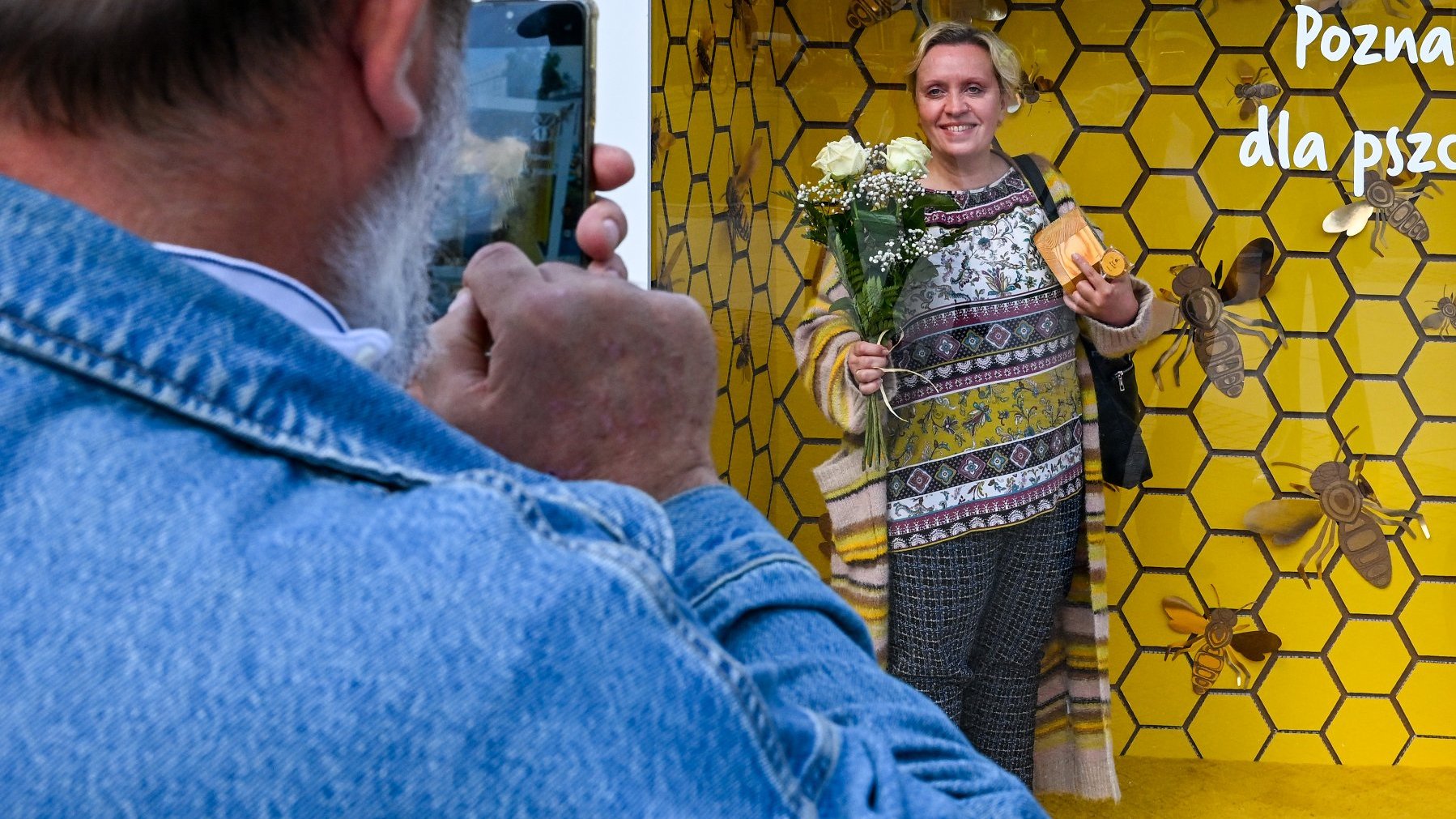 Zdjęcie przedstawia kobietę pozującą w instalacji imitującej ul z bukietem kwiatów oraz mężczyznę, który robi jej zdjęcie telefonem.