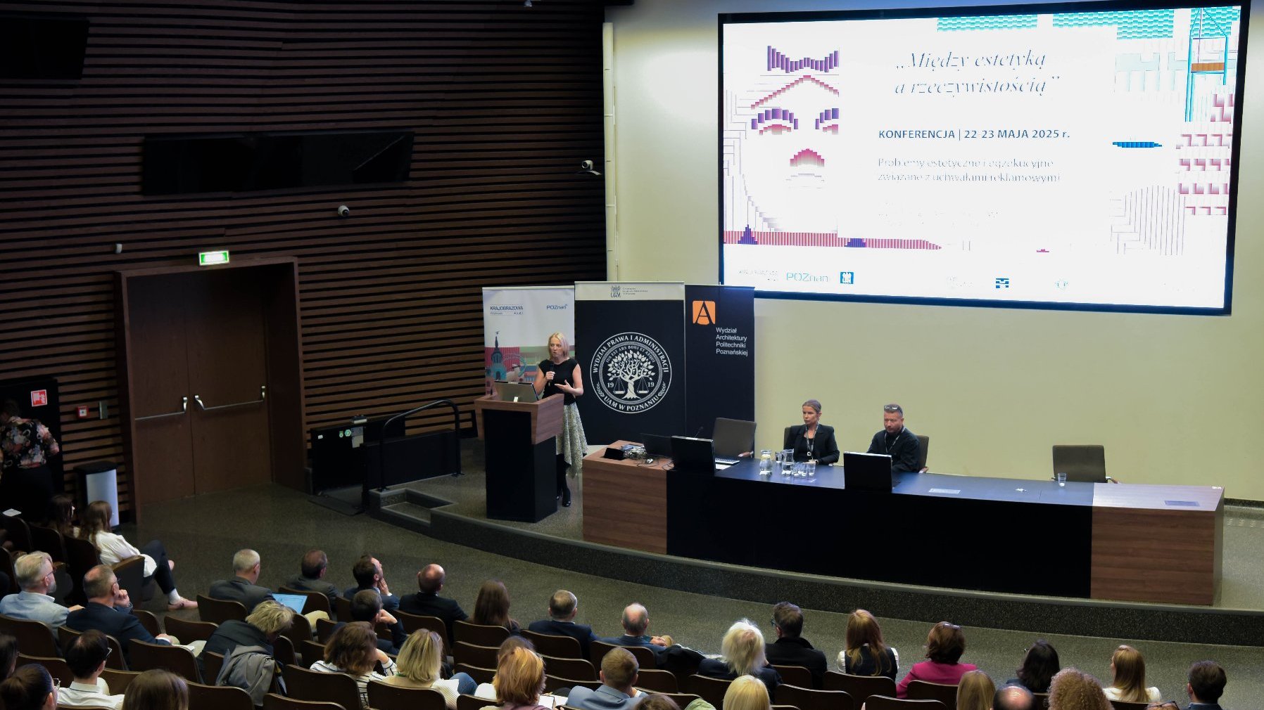 Konferencja "Między estetyką a rzeczywistością' odbywa się w dużej auli. Prelegent stoi przy podium, wygłaszając prezentację do zgromadzonej publiczności. Na ekranie za nim widnieje tytuł konferencji oraz szczegóły dotyczące terminu i tematyki.