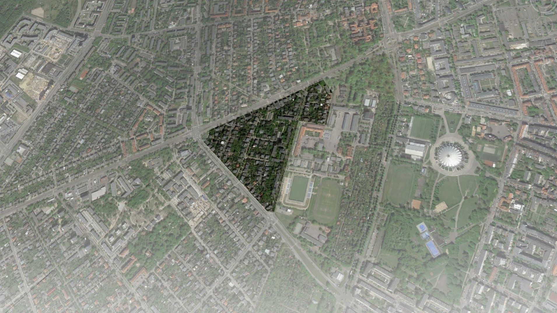 Widok z lotu ptaka na gęsto zabudowaną miejską przestrzeń. Widać mieszankę budynków mieszkalnych, komercyjnych i prawdopodobnie instytucjonalnych. Układ urbanistyczny charakteryzuje się siatką ulic i budynków, z kilkoma zielonymi przestrzeniami wplecionymi w krajobraz.