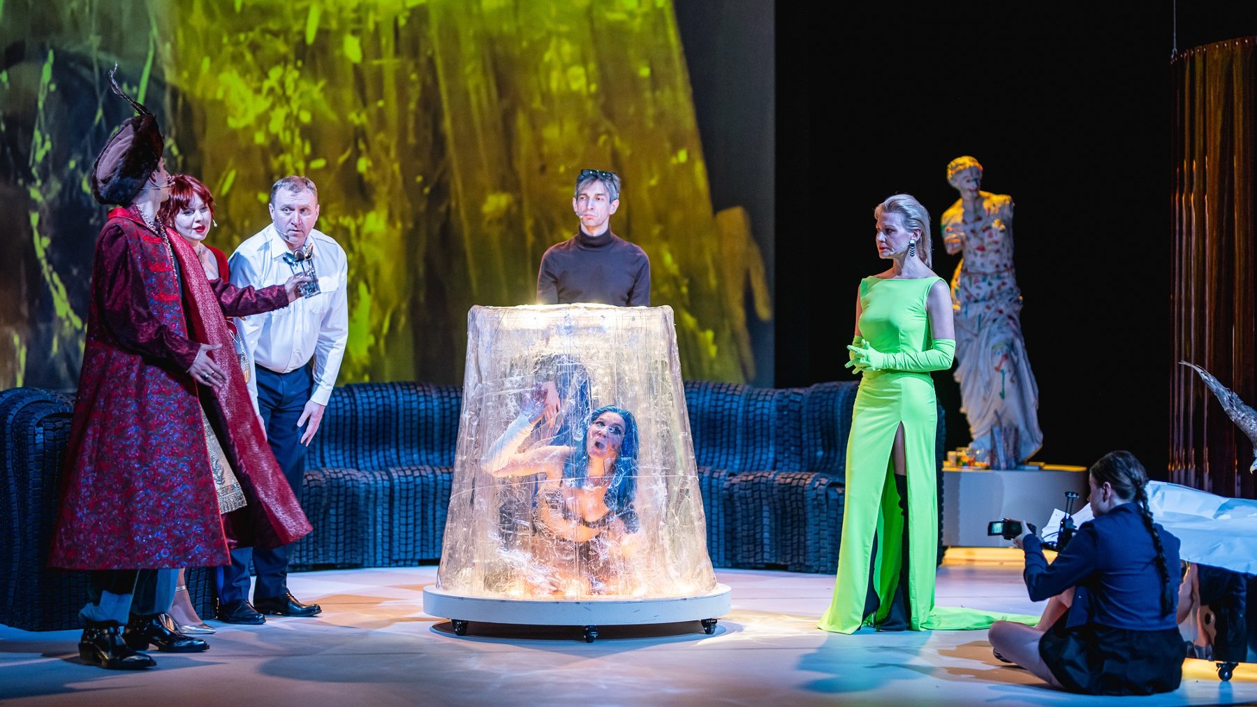 Scena teatralna z przedstawienia "Pigmalion". Pośrodku siedzi kobieta w niebieskiej peruce, otoczona przezroczystym, cylindrycznym kloszem. Dookoła niej stoją aktorzy w kostiumach. Po prawej kobieta w długiej, jasnozielonej sukni z rozcięciem.
