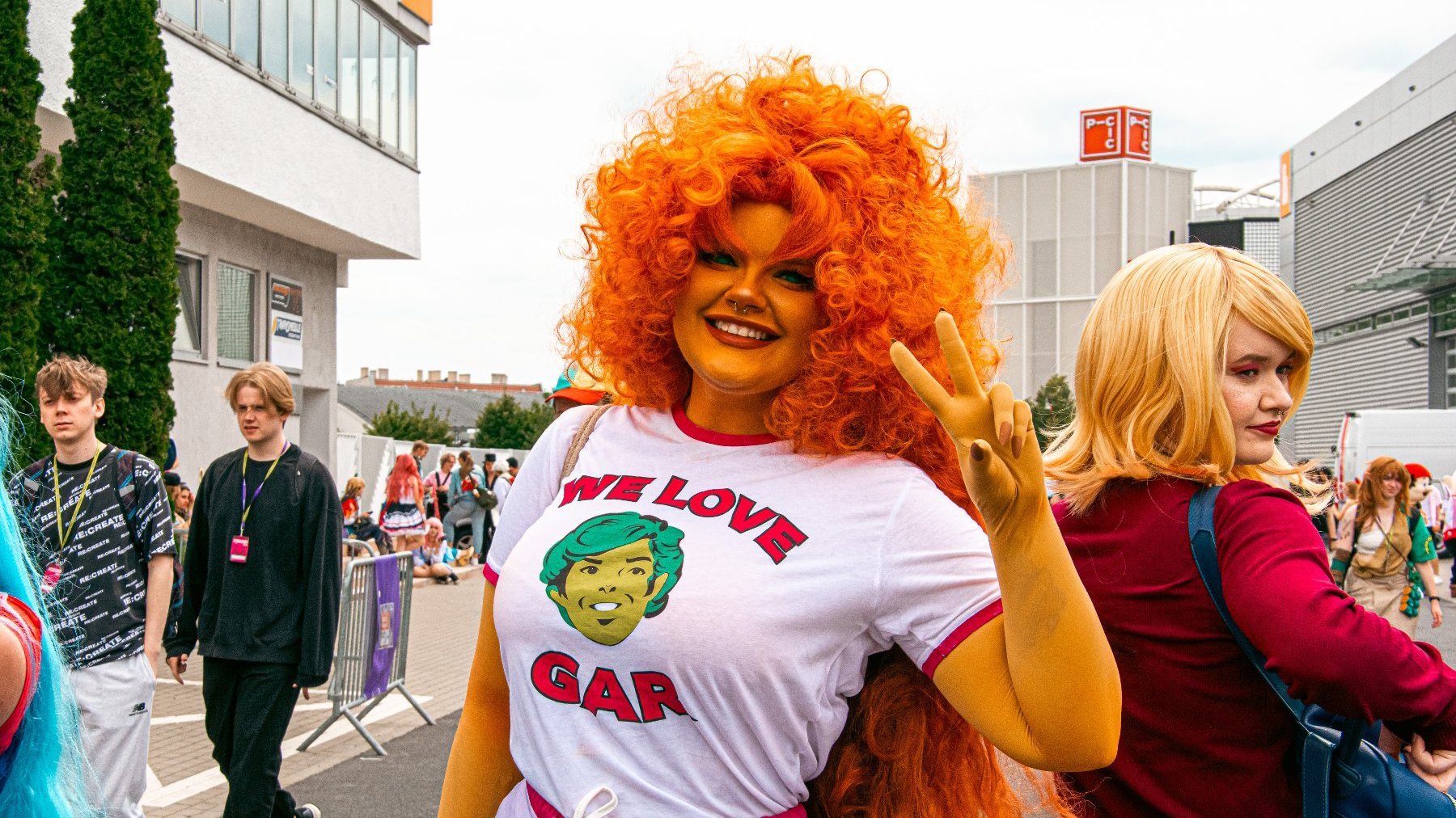 Zdjęcie przedstawia dziewczynę w peruce z burzą loków. Ma na sobie koszulkę z napisem "we love gar" i rysunkiem głowy mężczyzny. Obok widać tłum innych ludzi.