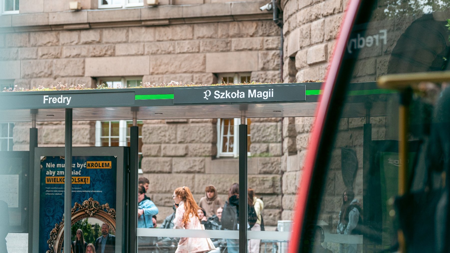 Zdjęcie przedstawia przystanek tramwajowy z napisem Szkoła Magii. Po chodniku chodzą ludzie.