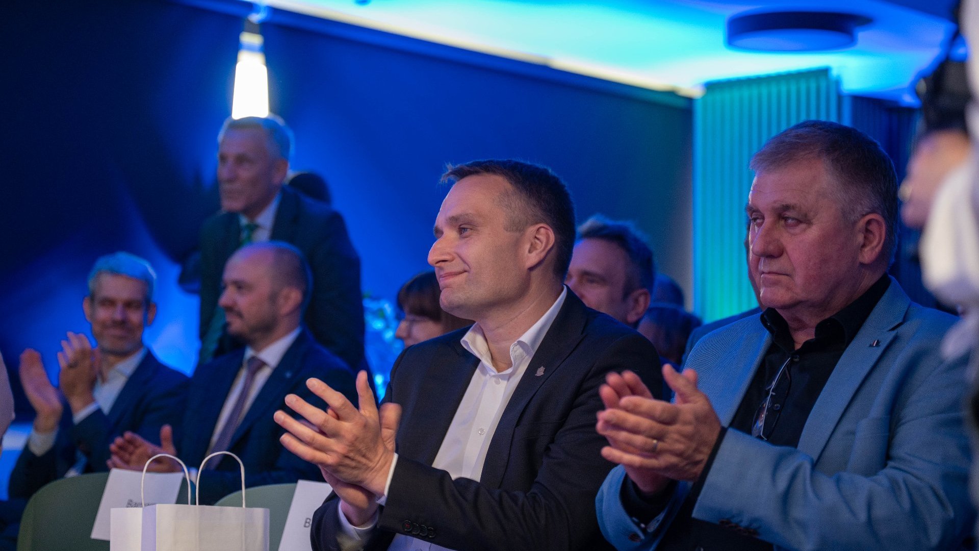 Grupa mężczyzn w eleganckich strojach siedzi i klaszcze, oświetlona niebieskim światłem. Po prawej stronie widać białką torbę z napisem.