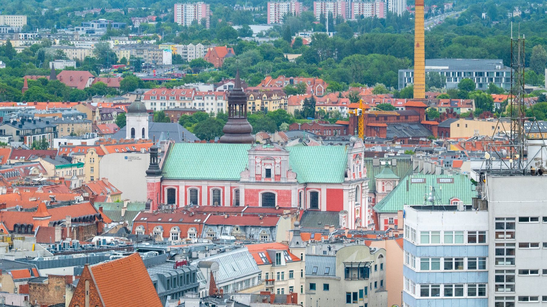 Widok na panoramę miasta Poznań, z licznymi budynkami mieszkalnymi i zabytkowymi. Na pierwszym planie wyróżnia się kościół o czerwono-białej fasadzie i zielonym dachu. W tle widać wieżę fabryczną.