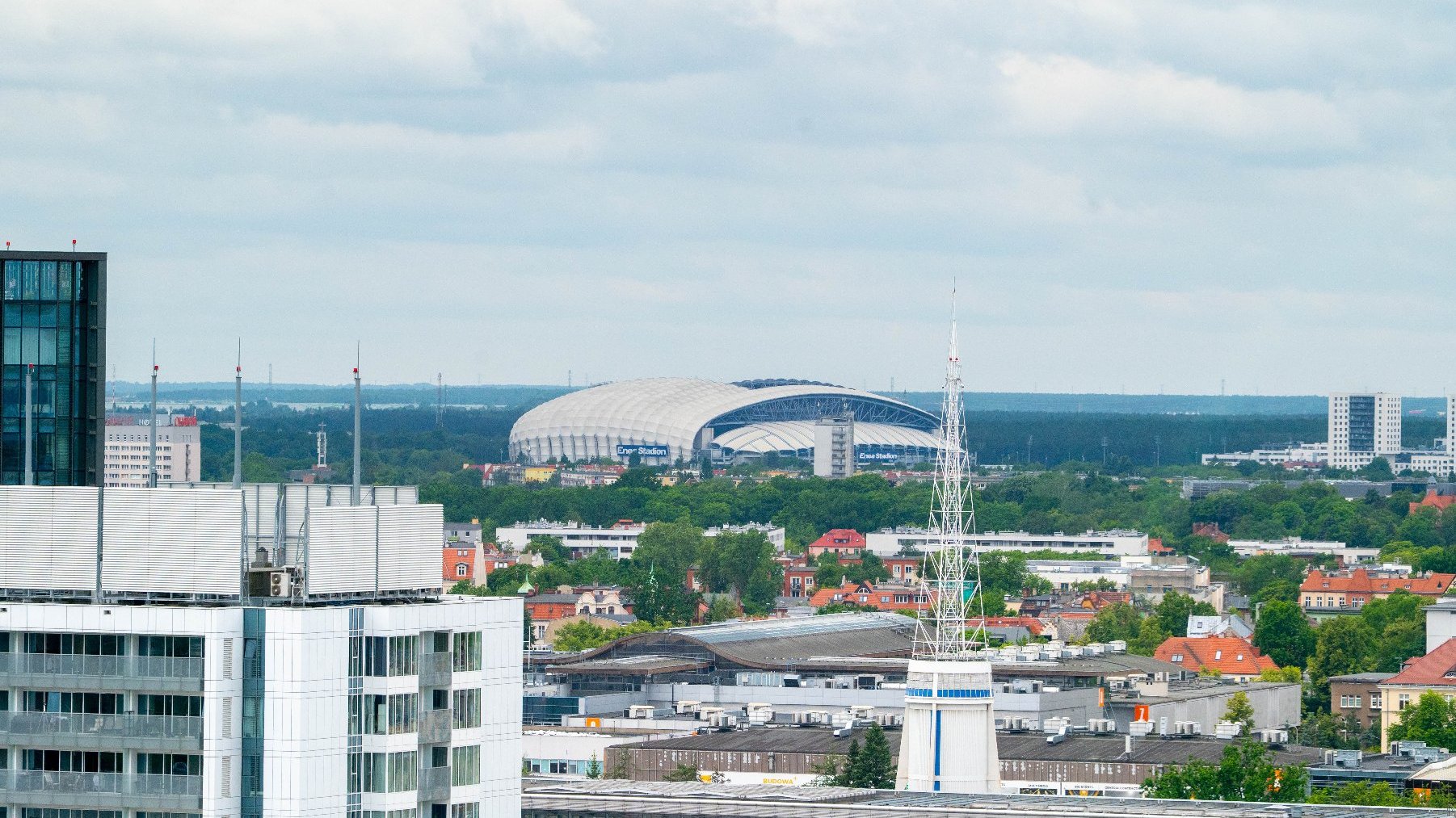 Panorama miasta z dominującym na horyzoncie dużym stadionem. Na pierwszym planie widoczne są dachy budynków mieszkalnych i biurowych, a także wysoka, ażurowa wieża.