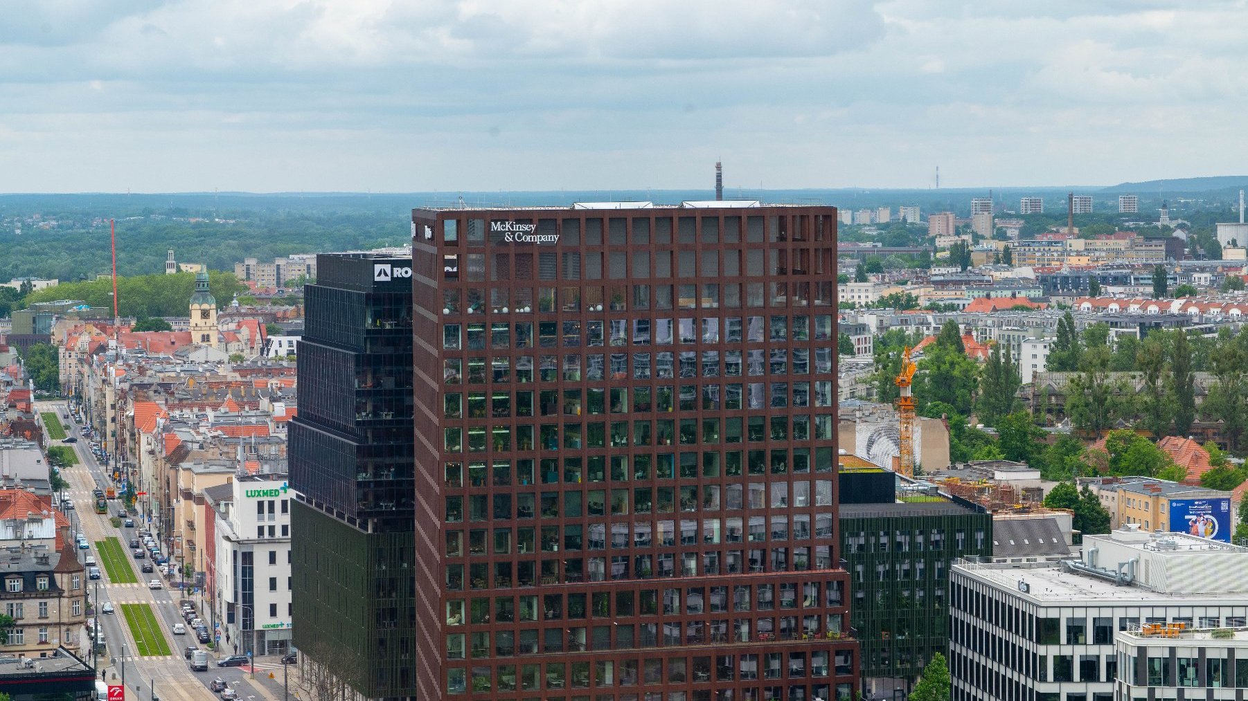 Widok na centrum Poznania z lotu ptaka. Na pierwszym planie widoczne są dwa nowoczesne wieżowce biurowe: jeden brązowy z napisem "McKinsey & Company", drugi ciemny, ze szklaną fasadą. W tle rozciąga się miejska zabudowa z ulicą i samochodami, a także liczne drzewa.