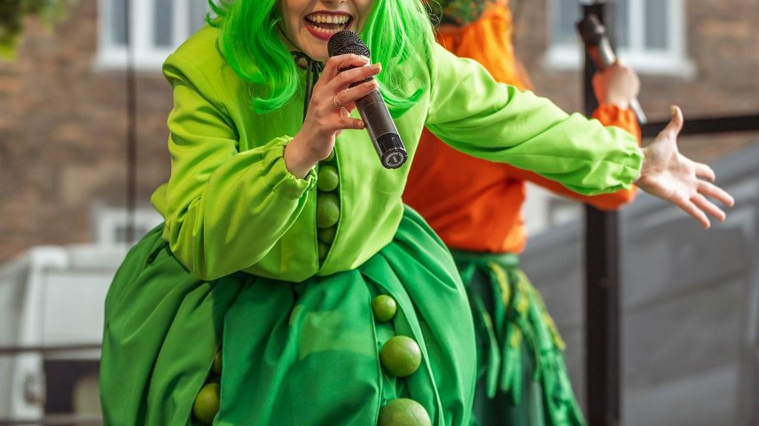 Zdjęcie przedstawia przebraną kobietę z mikrofonem na scenie.