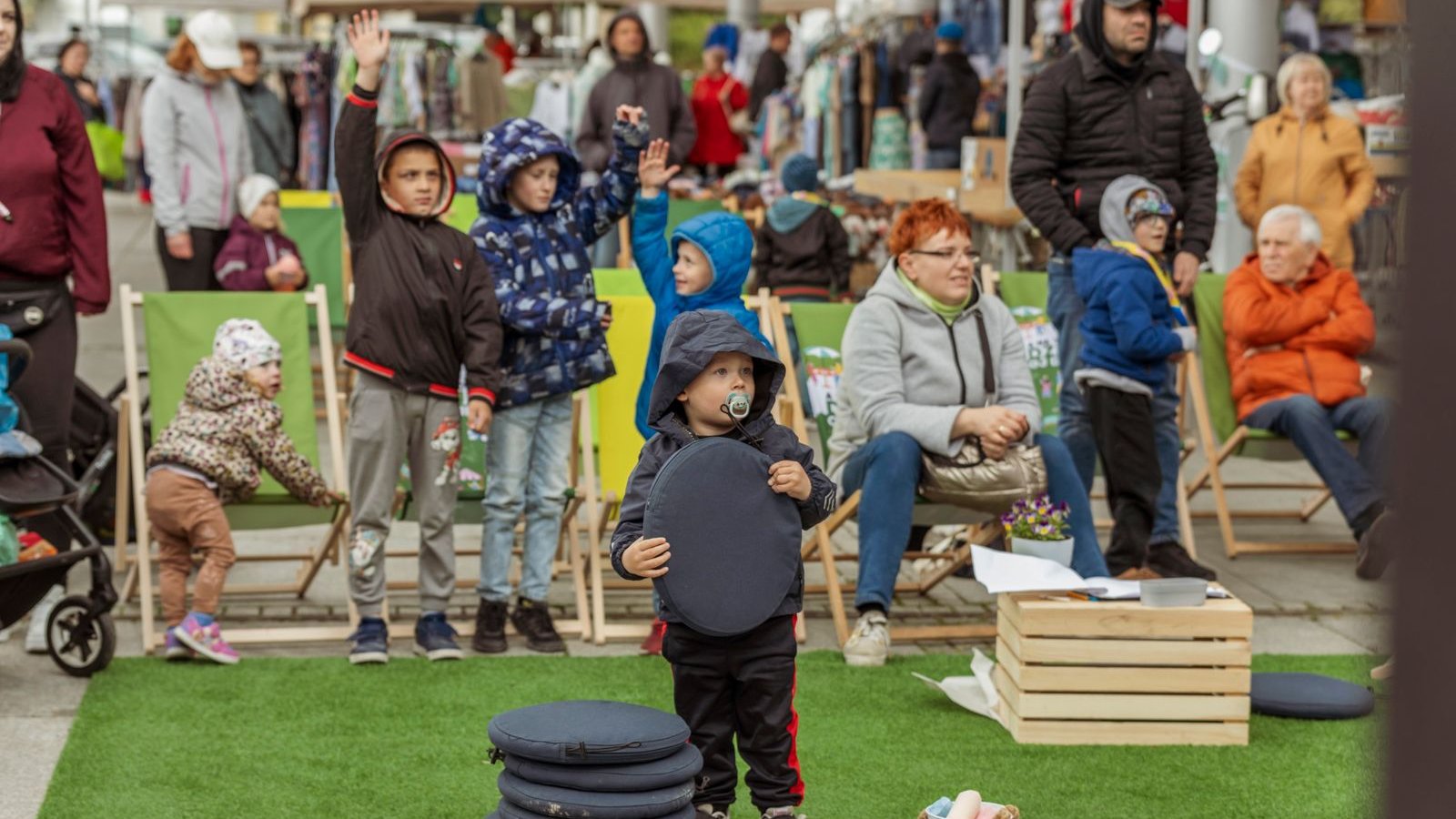 Zdjęcie przedstawia dzieci i dorosłych patrzących na jakieś wydarzenie. Niektóre dzieci podnoszą ręce w górę, niektórzy dorośli siedzą na leżakach.