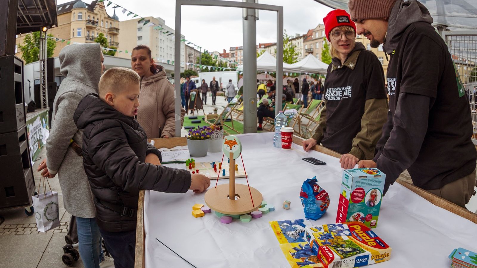 Zdjęcie przedstawia stół, na kórym leżą gry dla dzieci. Po jego jednej stronie stoi dwójka dorosłych, po drugiej - dziecko i dwie kobiety. W tle widać tłum ludzi i rozstawione leżaki.