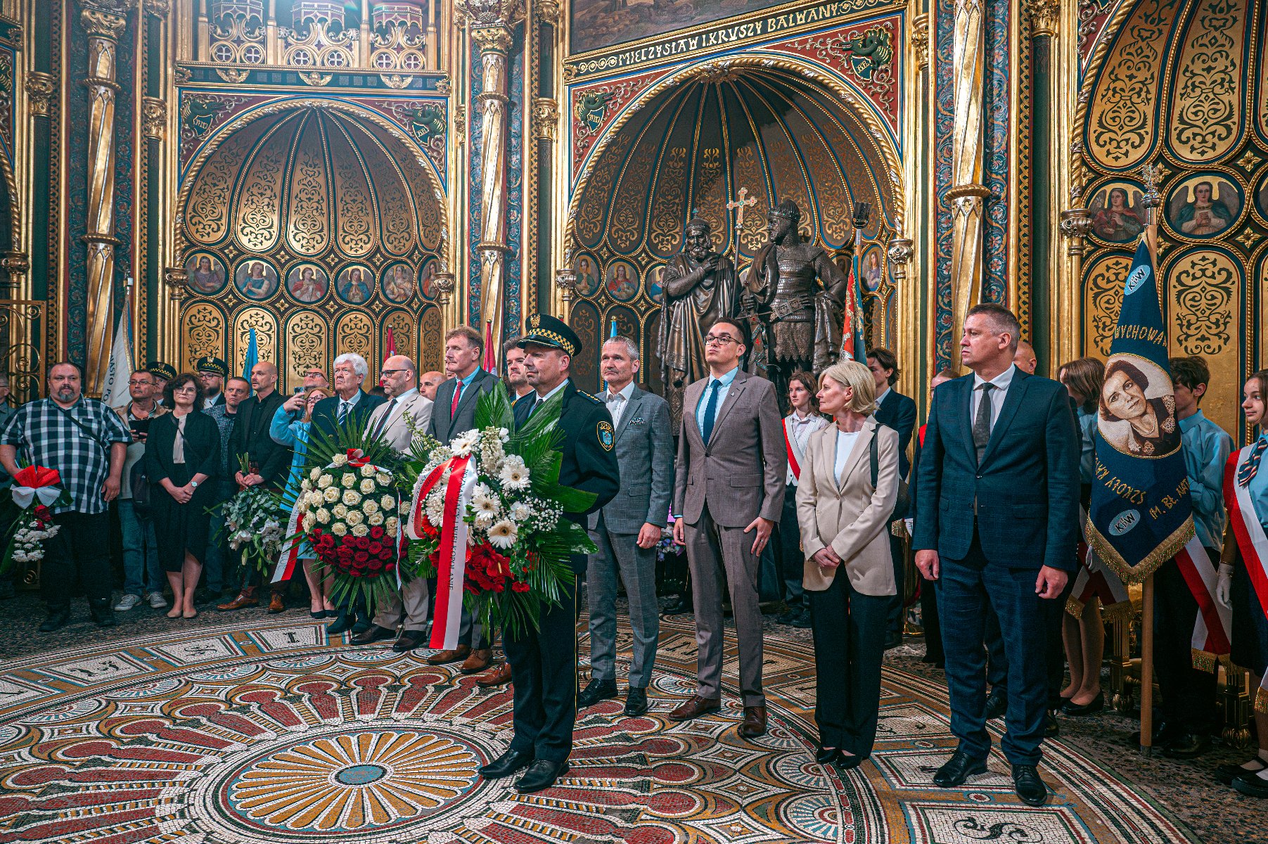 Zdjęcie przedstawia grupę ludzi w bogato ozdobionym pomieszczeniu. Przed nimi stoi strażnik miejski trzymający bukiet kwiatów. - grafika artykułu