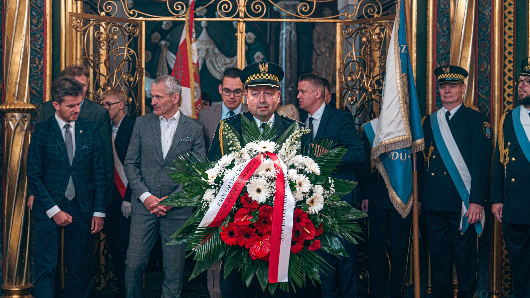 Zdjęcie przedstawia grupę ludzi w bogato ozdobionym pomieszczeniu. Przed nimi stoi strażnik miejski trzymający bukiet kwiatów.