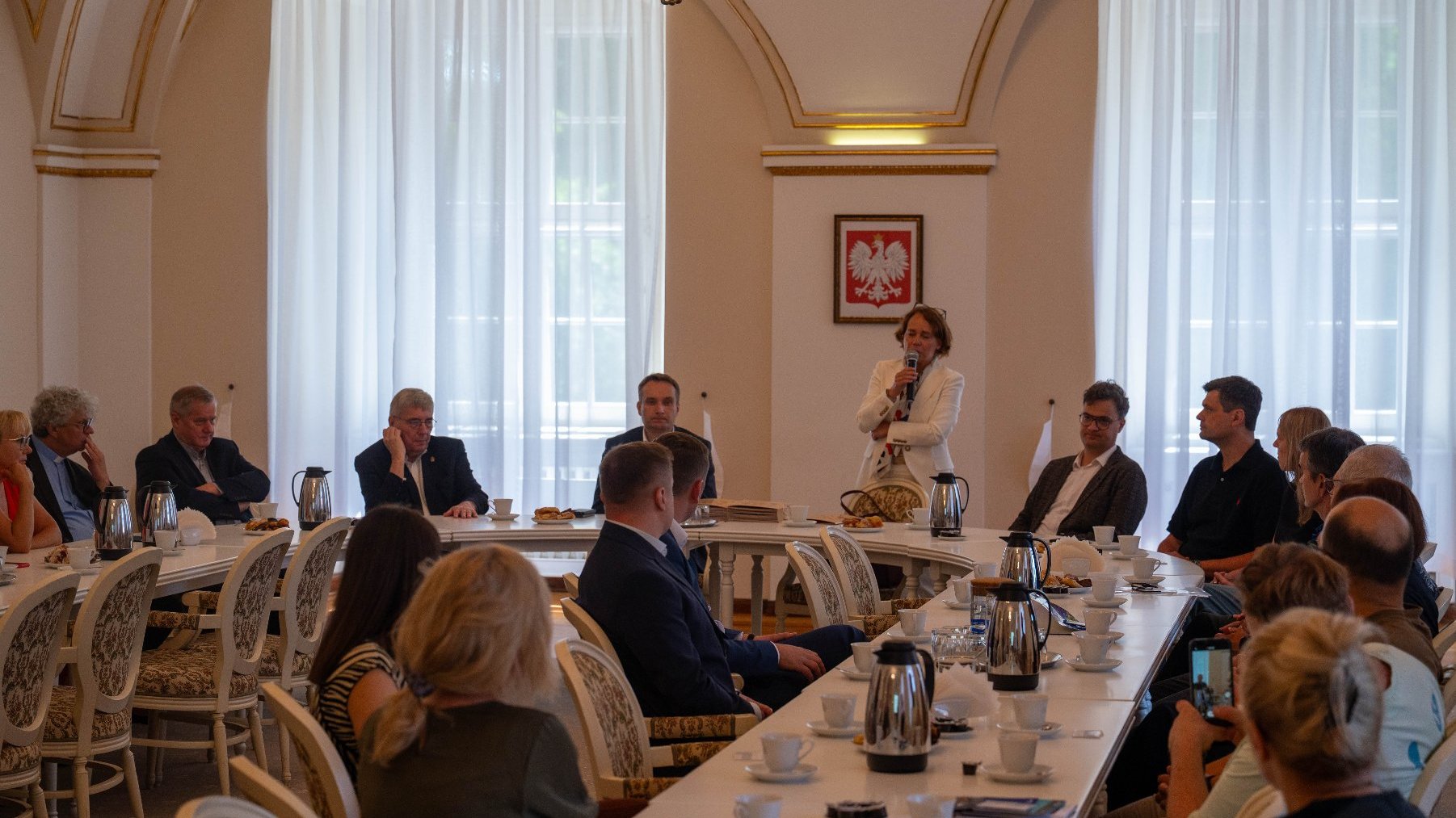Kobieta w białej marynarce przemawia do zebranych osób, trzymając mikrofon w dłoni. Spotkanie odbywa się w eleganckiej sali, przy dużym stole, na którym stoją filiżanki i dzbanki z kawą. W tle widoczne są duże okna z białymi zasłonami oraz obraz z polskim godłem.