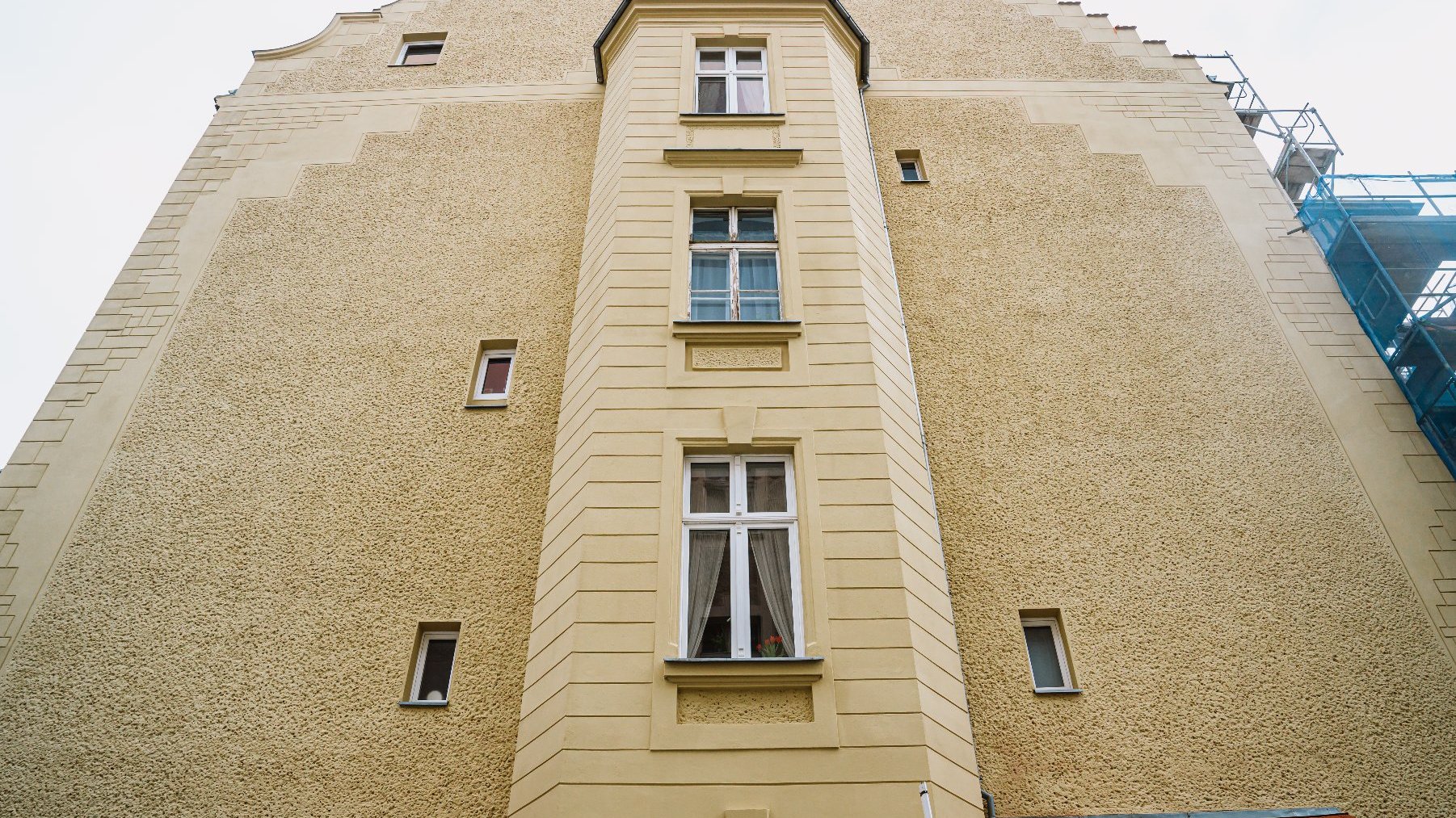 Zabytkowa kamienica w Poznaniu, z widokiem od dołu. Budynek ma beżową elewację z wyraźną fakturą. Na środku znajduje się prostokątny wykusz z trzema oknami, otoczony gładkim, jaśniejszym tynkiem.