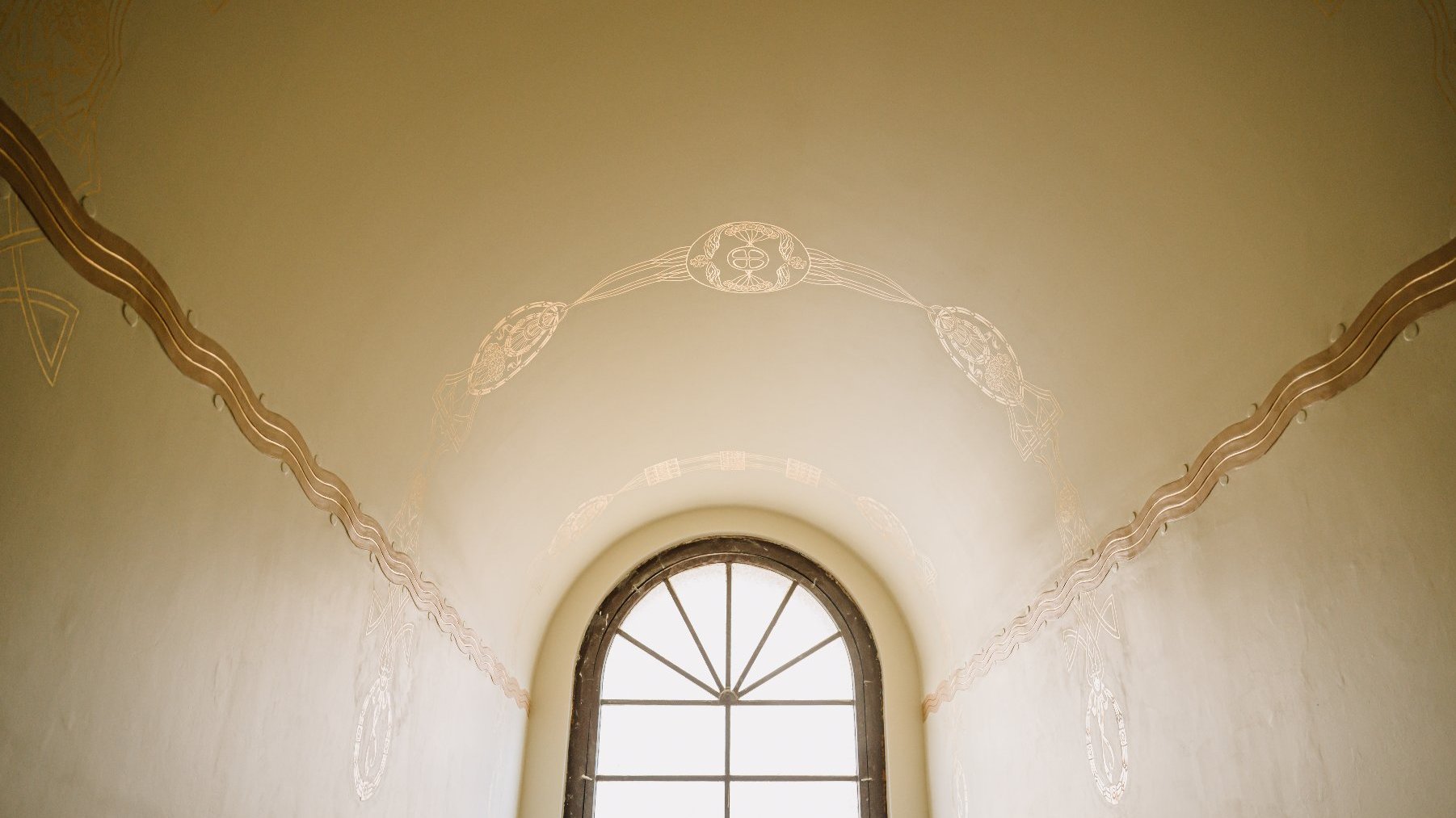 Widok od wewnątrz na półokrągłe okno, przez które wpada dzienne światło. Wokół okna ściany i sufit zdobione jasnymi i złotymi wzorami. Po prawej i lewej stronie widoczne złote, falujące linie. Nad oknem złota ozdoba w kształcie łuku.