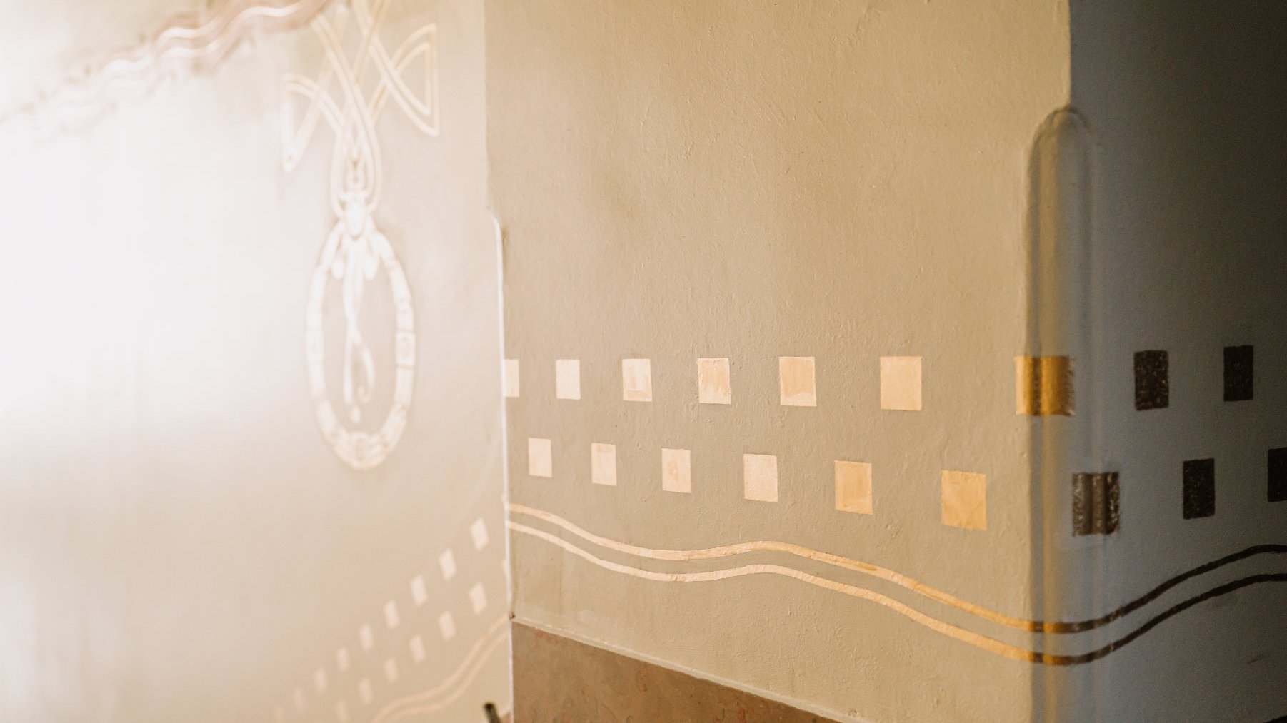 Narożnik jasnych ścian wewnętrznych z ozdobnymi wzorami. Na prawej ścianie widoczne są złote kwadraty ułożone w rzędy oraz dwie faliste, złote linie poniżej. Po lewej stronie, na ścianie w tle, znajduje się duży, owalny, jasny ornament z krzyżującymi się liniami. Na dole fragment brązowego cokołu.