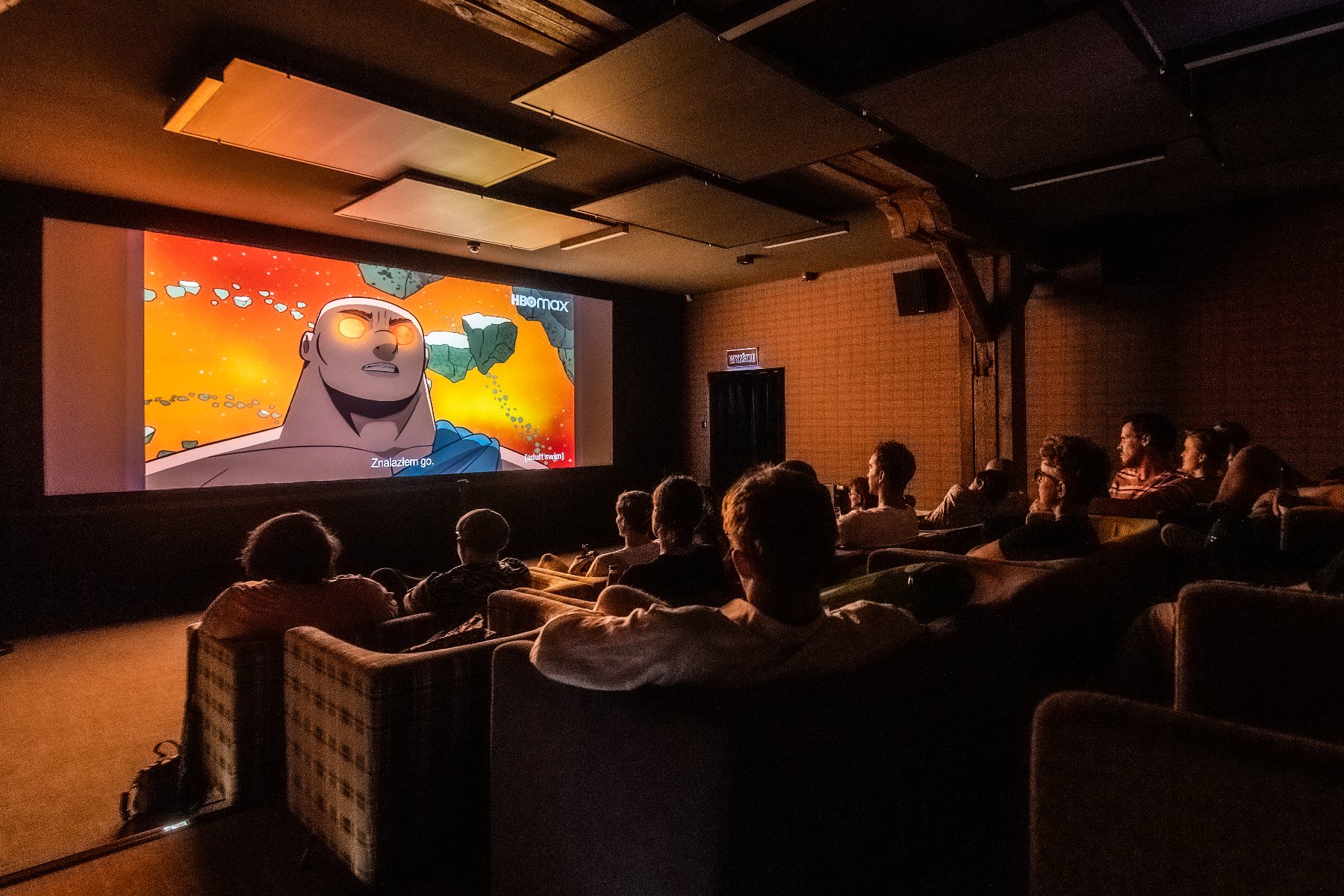 Kino studyjne w Poznaniu. Widownia pełna ludzi oglądających film animowany. Na ekranie widać postać z kreskówki i napis "HBO MAX". Sala jest ciemna, a światło pada z ekranu. Sufit ozdobiony jest prostokątnymi panelami. - grafika artykułu