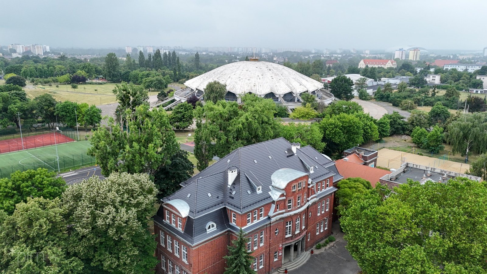 Na zdjęciu zabytkowy budynek z nowym dachem, widziany z lotu ptaka, w tle Arena