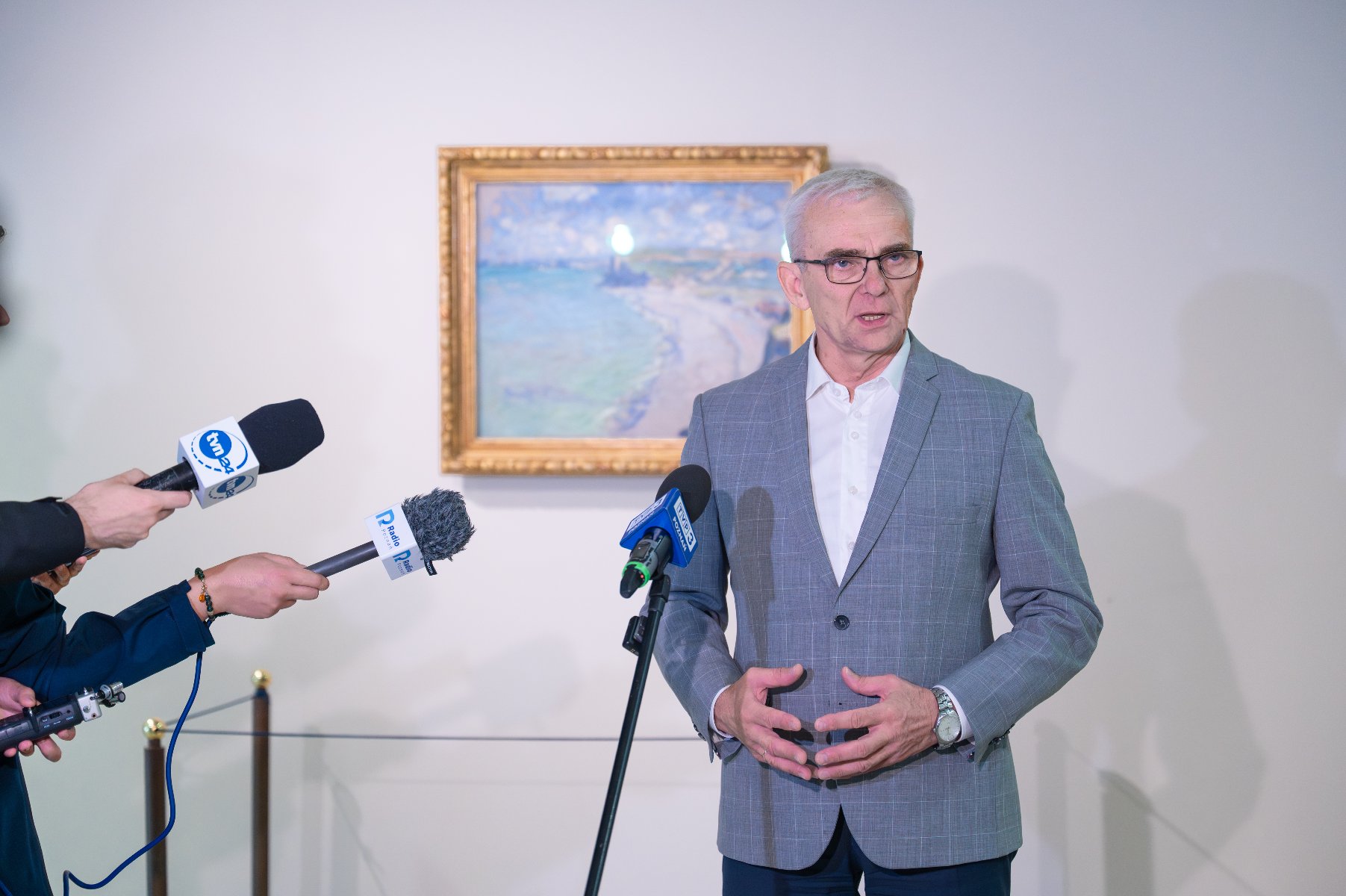 Na zdjęciu mężczyzna w garniturze mówi coś do mikrofonu, za nim wisi obraz, obok widać ręce dziennikarzy radiowych trzymających mikrofony - grafika artykułu