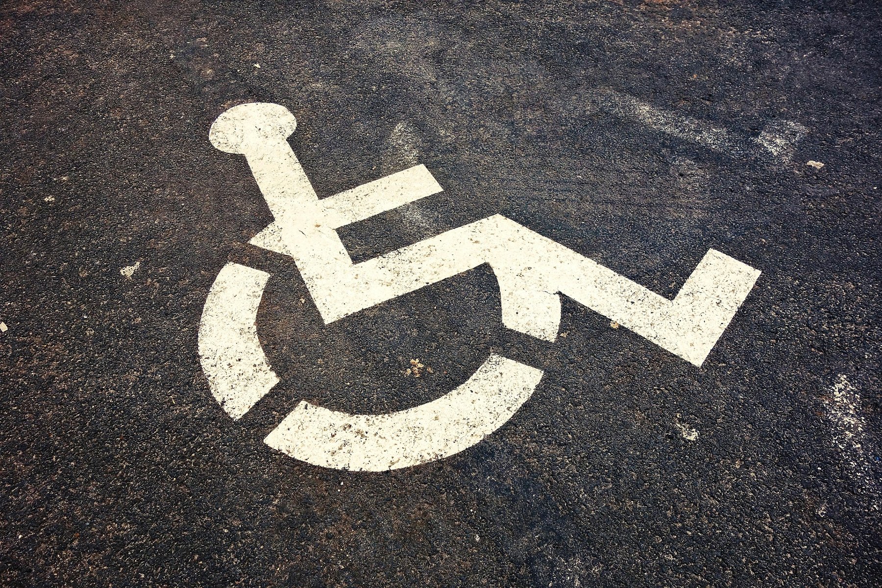 Biały piktogram wózka inwalidzkiego, symbolizujący miejsce parkingowe dla osób z niepełnosprawnościami, namalowany na ciemnym asfalcie. - grafika artykułu