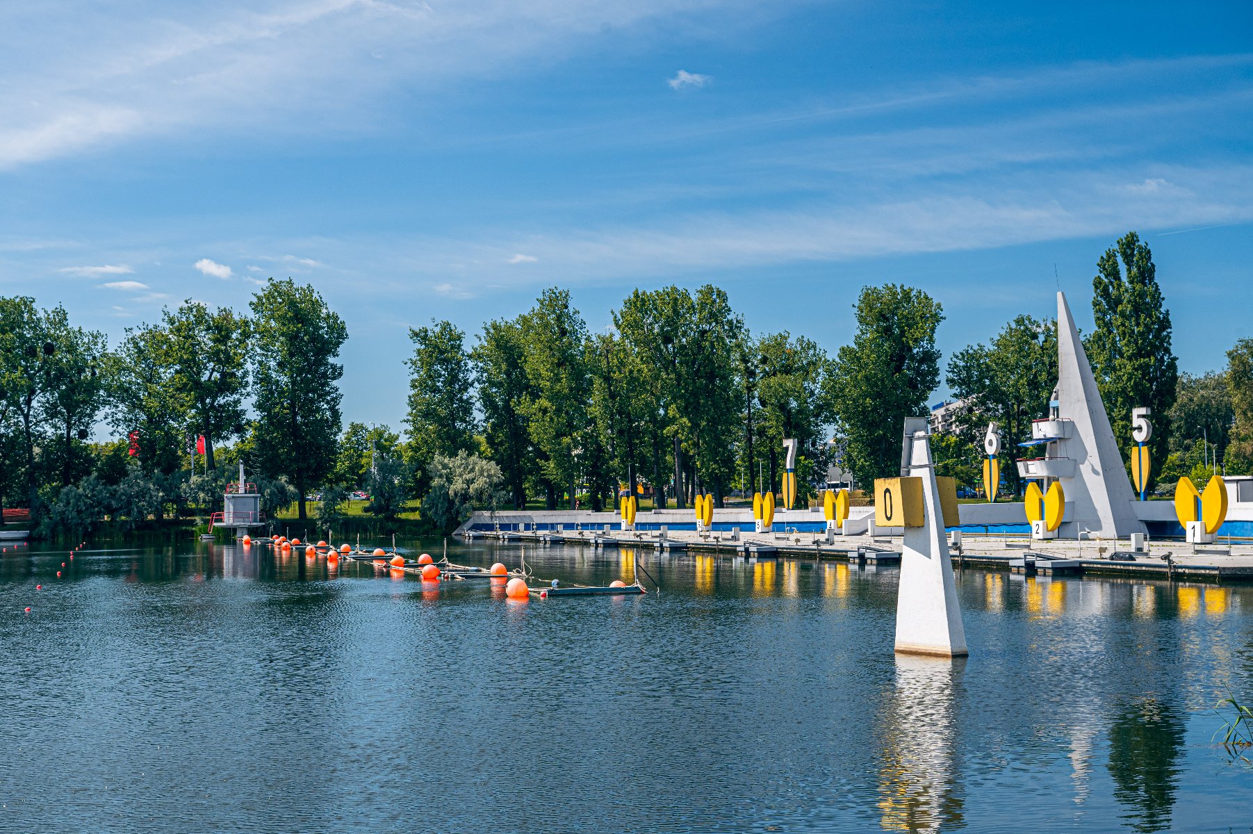 or regatowy Malta w Poznaniu, z widoczną przystanią, bojkami na wodzie i metą z numerami stanowisk. W tle zielone drzewa i błękitne niebo z kilkoma chmurami. - grafika artykułu