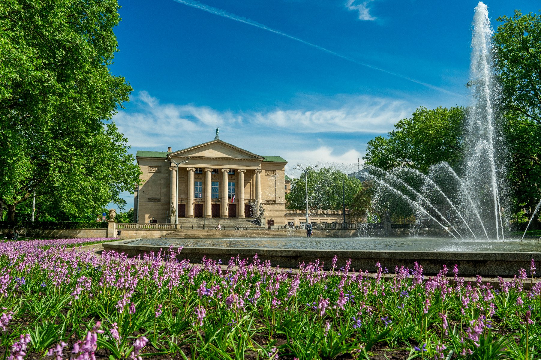 Fasada Teatru Wielkiego im. Stanisława Moniuszki w Poznaniu, otoczonego zielonym parkiem z kwitnącymi, liliowymi kwiatami na pierwszym planie. Przed teatrem znajduje się duża fontanna tryskająca wodą. Błękitne niebo z lekkimi smugami chmur dopełnia obraz. - grafika artykułu