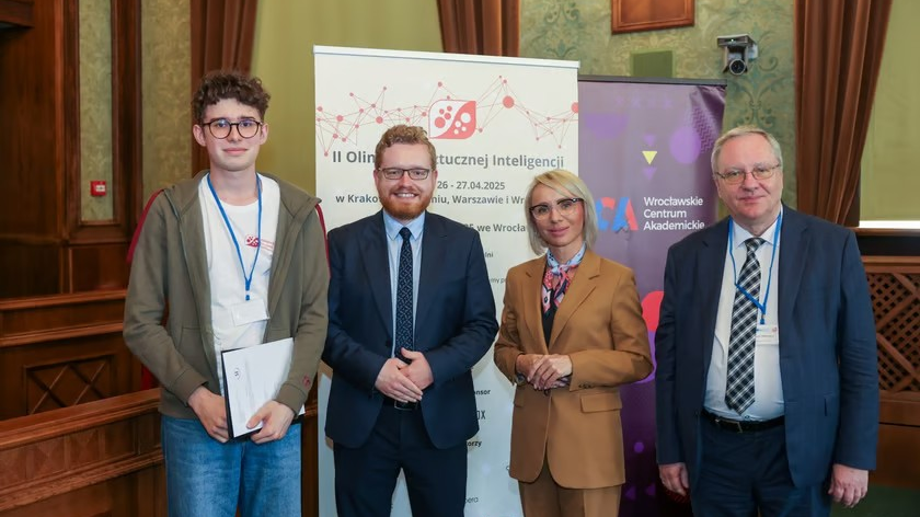 Cztery osoby stojące obok siebie na tle banera informującego o II Olimpiadzie Sztucznej Inteligencji. Od lewej: młody mężczyzna w okularach i jasnej bluzie z identyfikatorem na szyi, uśmiechnięty mężczyzna w garniturze z krawatem, kobieta w brązowym garniturze z chustą na szyi i okularach oraz starszy mężczyzna w garniturze z krawatem i okularach. W tle po prawej widoczne jest logo Wrocławskiego Centrum Akademickiego. - grafika artykułu