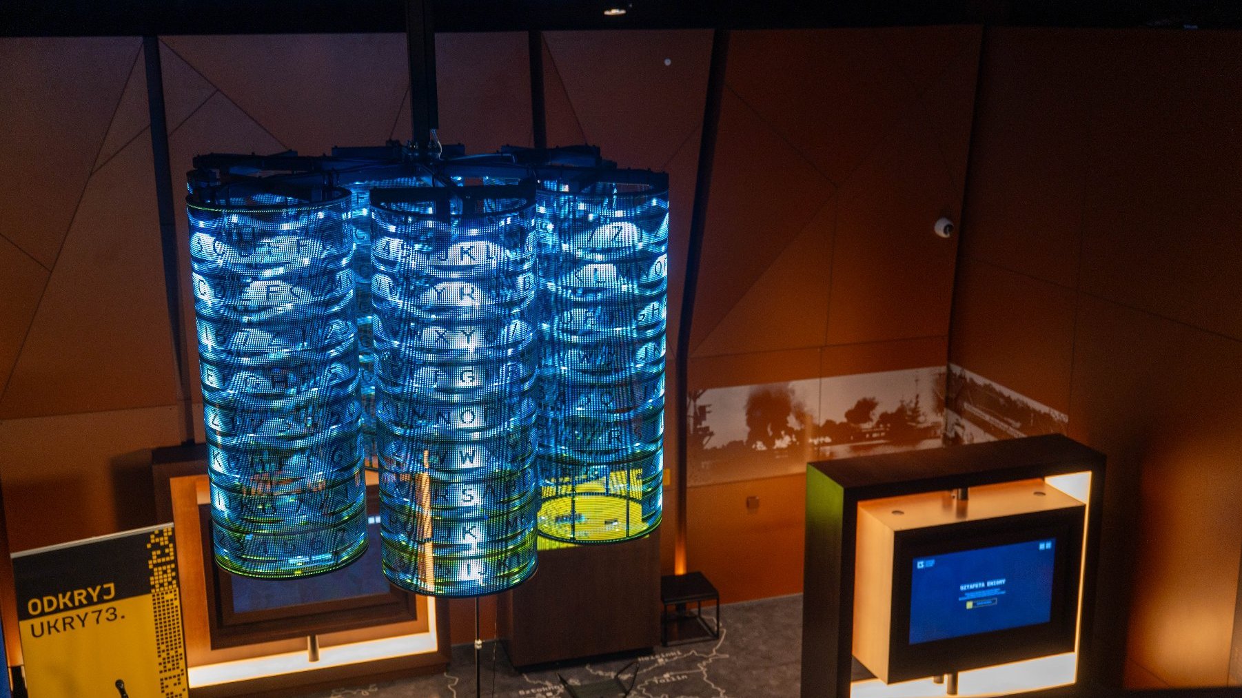 Nowoczesna instalacja artystyczna w kształcie trzech walców, świecąca na niebiesko i wyświetlająca litery, przypominająca szyfrowanie. Instalacja wisi w pomieszczeniu z brązowymi ścianami, a na podłodze widoczna jest mapa.