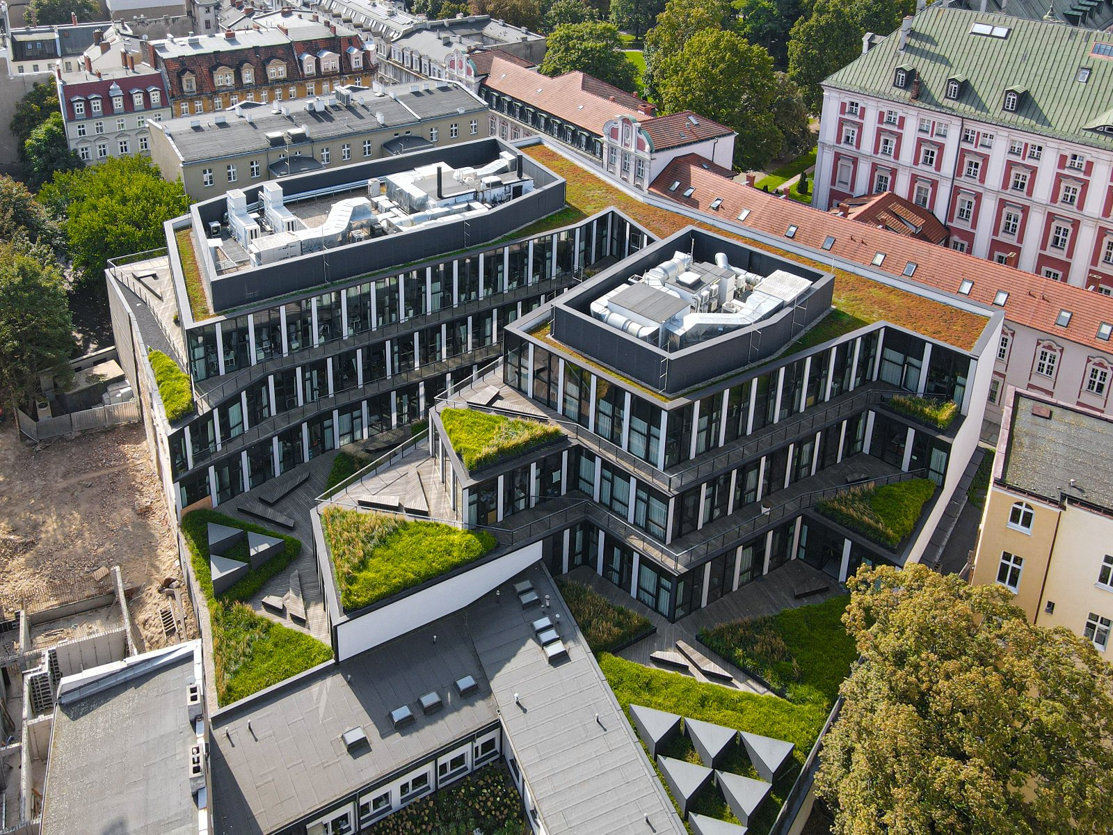 Widok z lotu ptaka na nowoczesny, wielopoziomowy budynek o szarych elewacjach. Na dachach i tarasach znajdują się liczne ogrody i trawniki. W tle widać starsze kamienice i drzewa. - grafika artykułu
