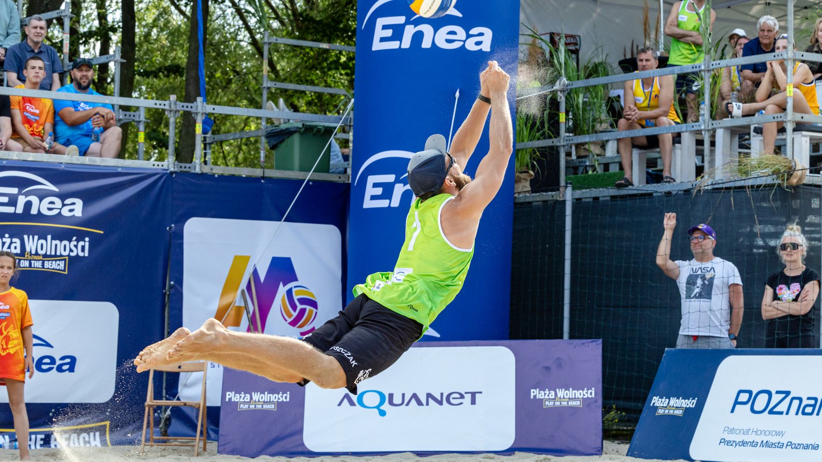 Siatkarz plażowy w zielonej koszulce i czarnych spodenkach skacze w powietrzu, przygotowując się do ataku. W tle widoczny jest niebieski baner sponsora, trybuny z kibicami i piasek. Na banerach widoczne są loga, a na jednym z nich napis "Poznań".