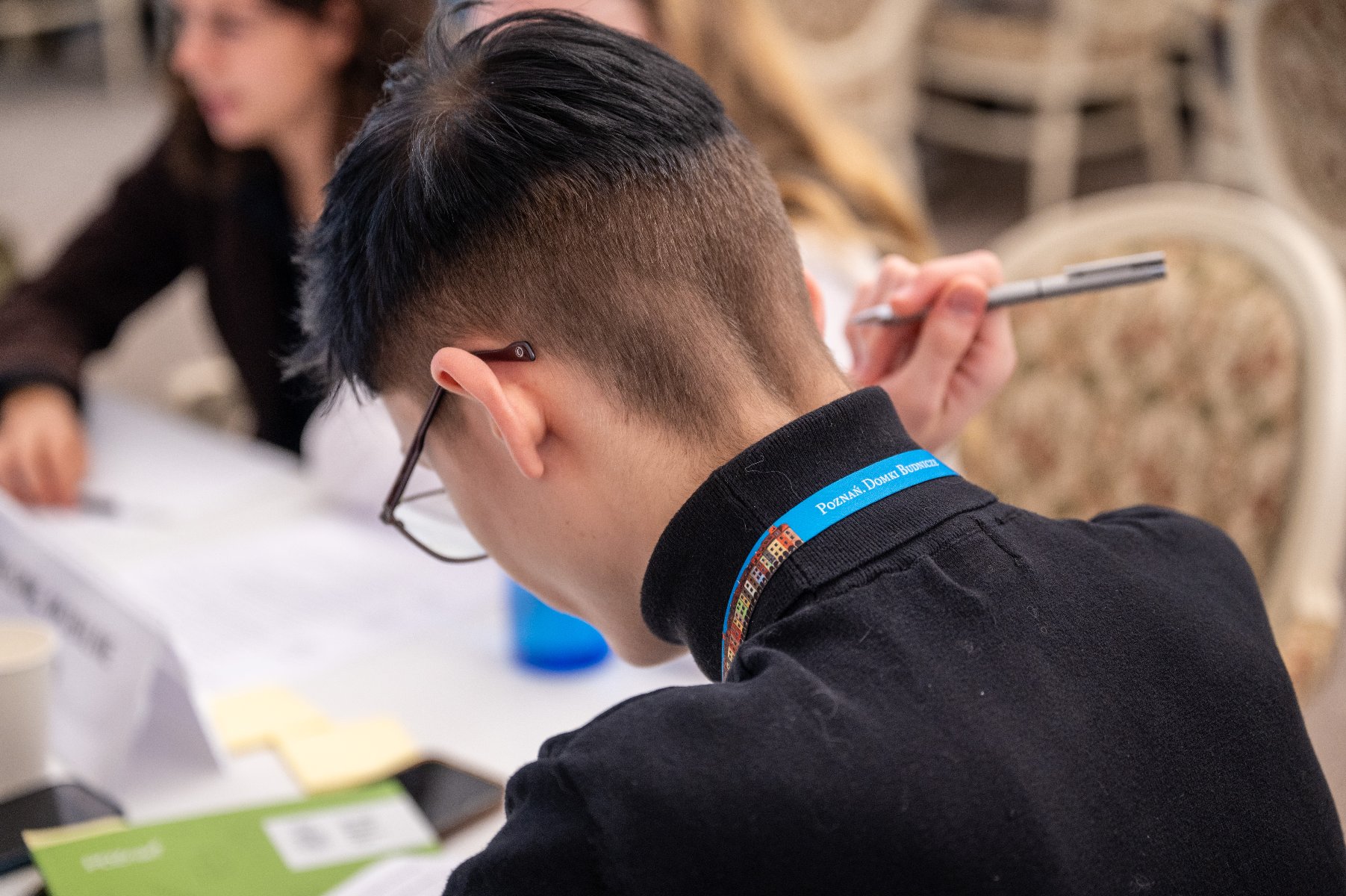Zdjęcie zrobione z tyłu, przedstawiające studenta z krótko ściętymi, czarnymi włosami, pochylającego się nad notatkami. Na kołnierzu ma smycz z napisem "Poznan' Dobre Biznesy". Obok niego siedzi inna osoba, trzymająca w ręku długopis. - grafika artykułu