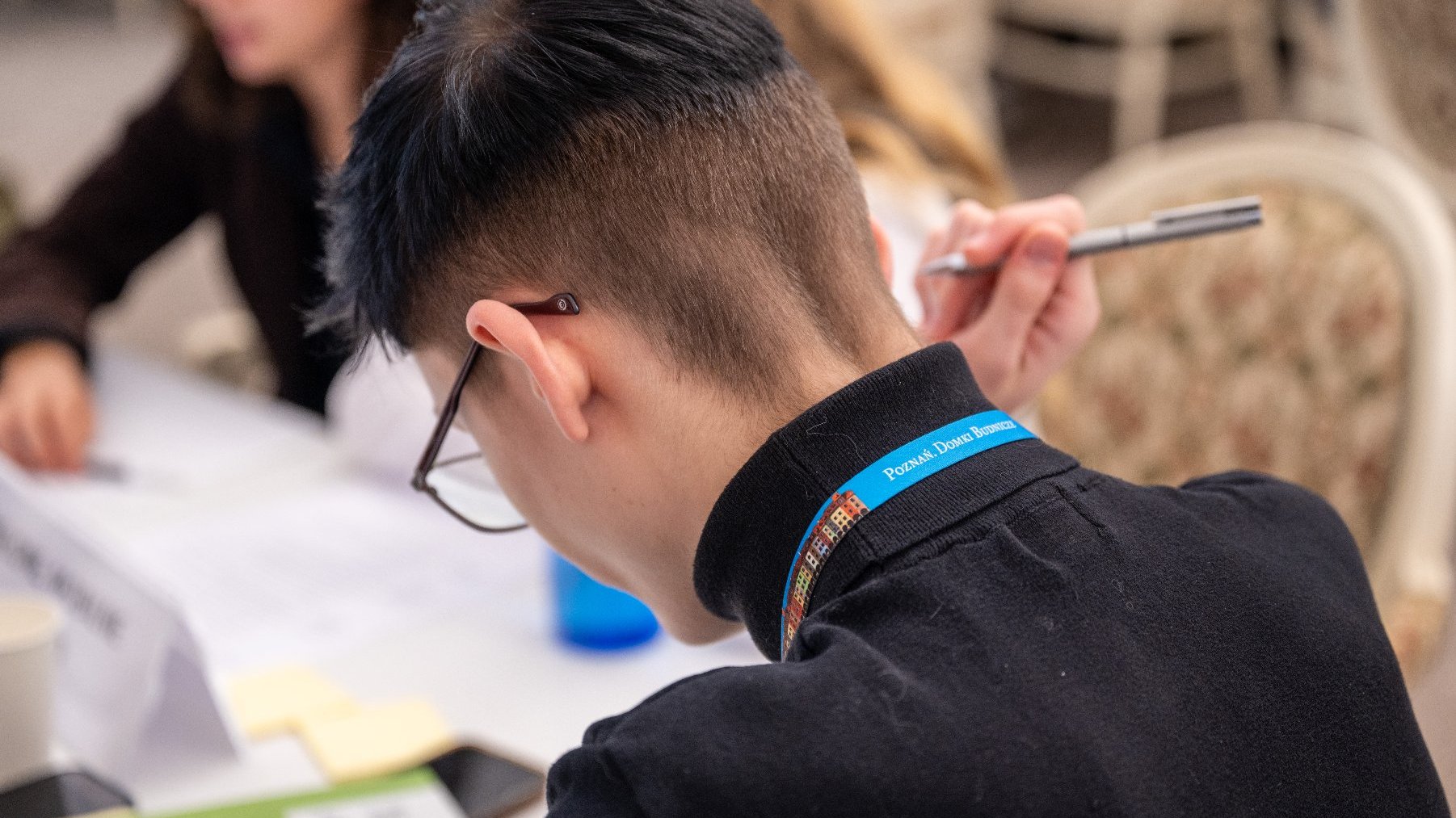 Zdjęcie zrobione z tyłu, przedstawiające studenta z krótko ściętymi, czarnymi włosami, pochylającego się nad notatkami. Na kołnierzu ma smycz z napisem "Poznan' Dobre Biznesy". Obok niego siedzi inna osoba, trzymająca w ręku długopis.