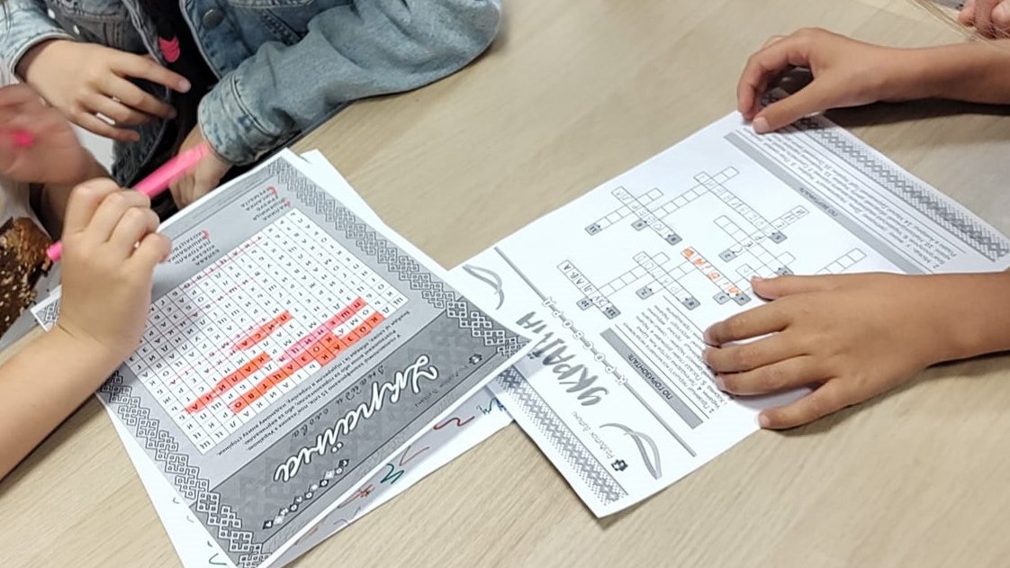 Dzieci siedzą przy stole i rozwiązują łamigłówki z napisami w języku ukraińskim. Na stole leży krzyżówka i wykreślanka.