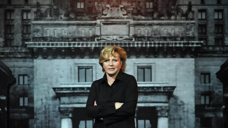 Na zdjęciu Krystyna Janda - aktorka w czarnej koszuli pozuje na tle zdjęcia kamienicy