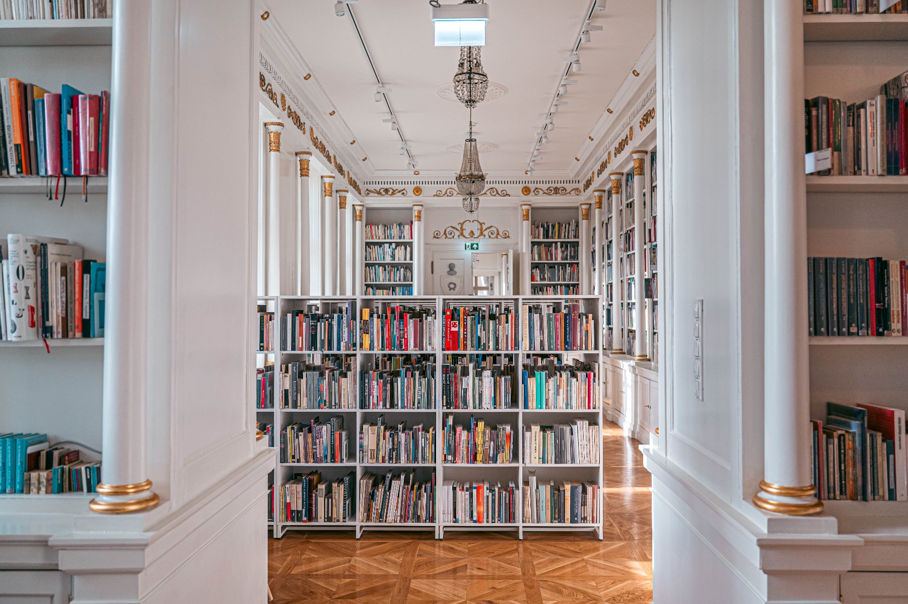 Eleganckie wnętrze biblioteki, pełne białych regałów z książkami. Na podłodze widać drewniany parkiet. Na suficie wiszą żyrandole, a na ścianach są złote detale. - grafika artykułu