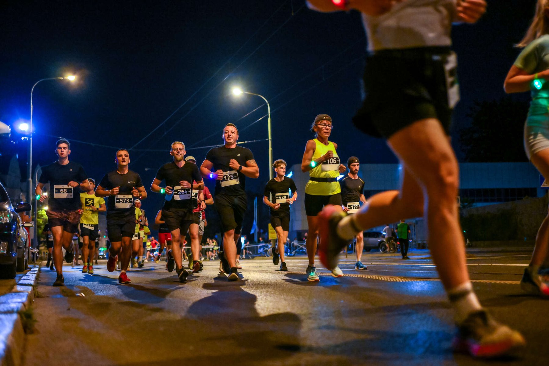 Wieczorny bieg uliczny, w którym biorą udział liczni zawodnicy. Wszyscy ubrani są w sportowe stroje, a niektórzy mają numery startowe. Mężczyzna w czarnej koszulce z numerem 1251 uśmiecha się. Po bokach drogi stoją latarnie uliczne, a w tle widać samochody. - grafika artykułu