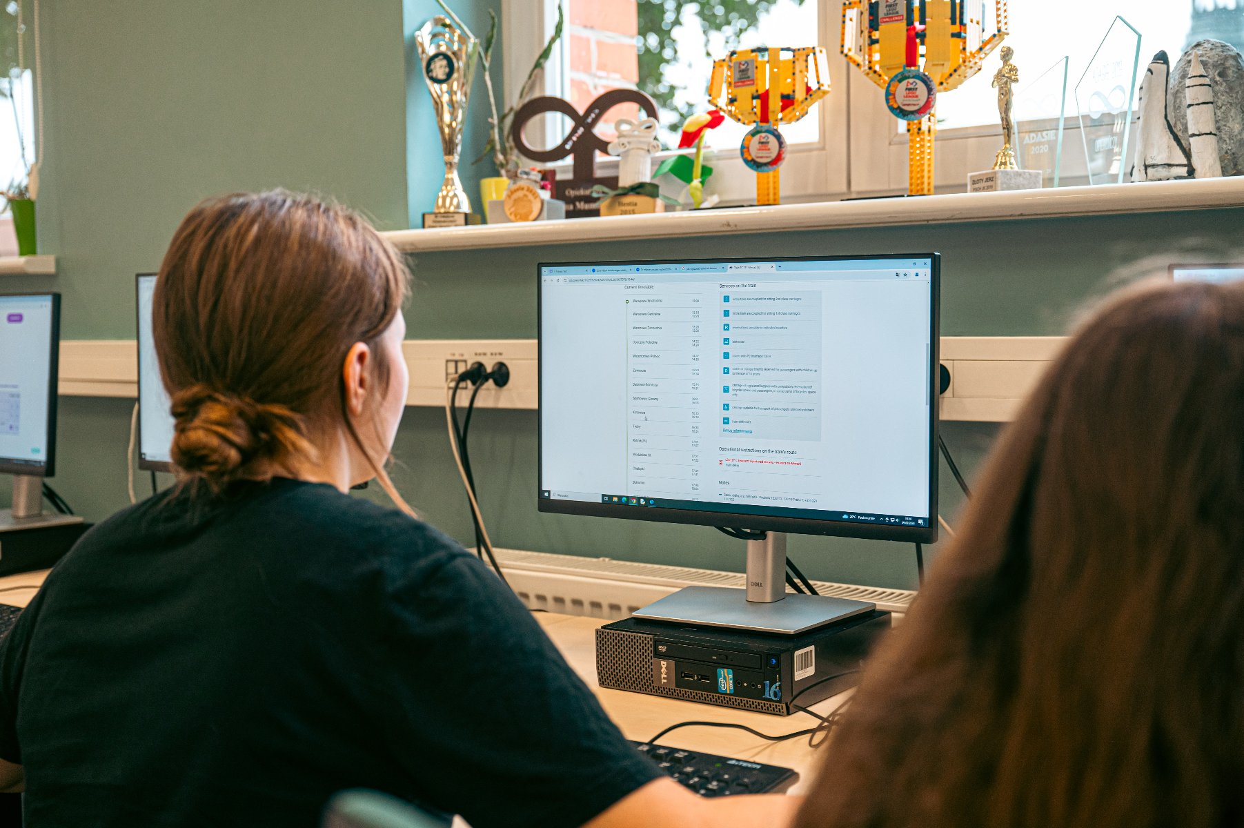 Dziewczyna siedzi tyłem do kamery przed monitorem komputerowym, pracując. Na ekranie widać otwartą aplikację. Na biurku obok komputera leży klawiatura. W tle, na parapecie, stoją liczne puchary i trofea, w tym model promu kosmicznego i statuetka robota z klocków. - grafika artykułu