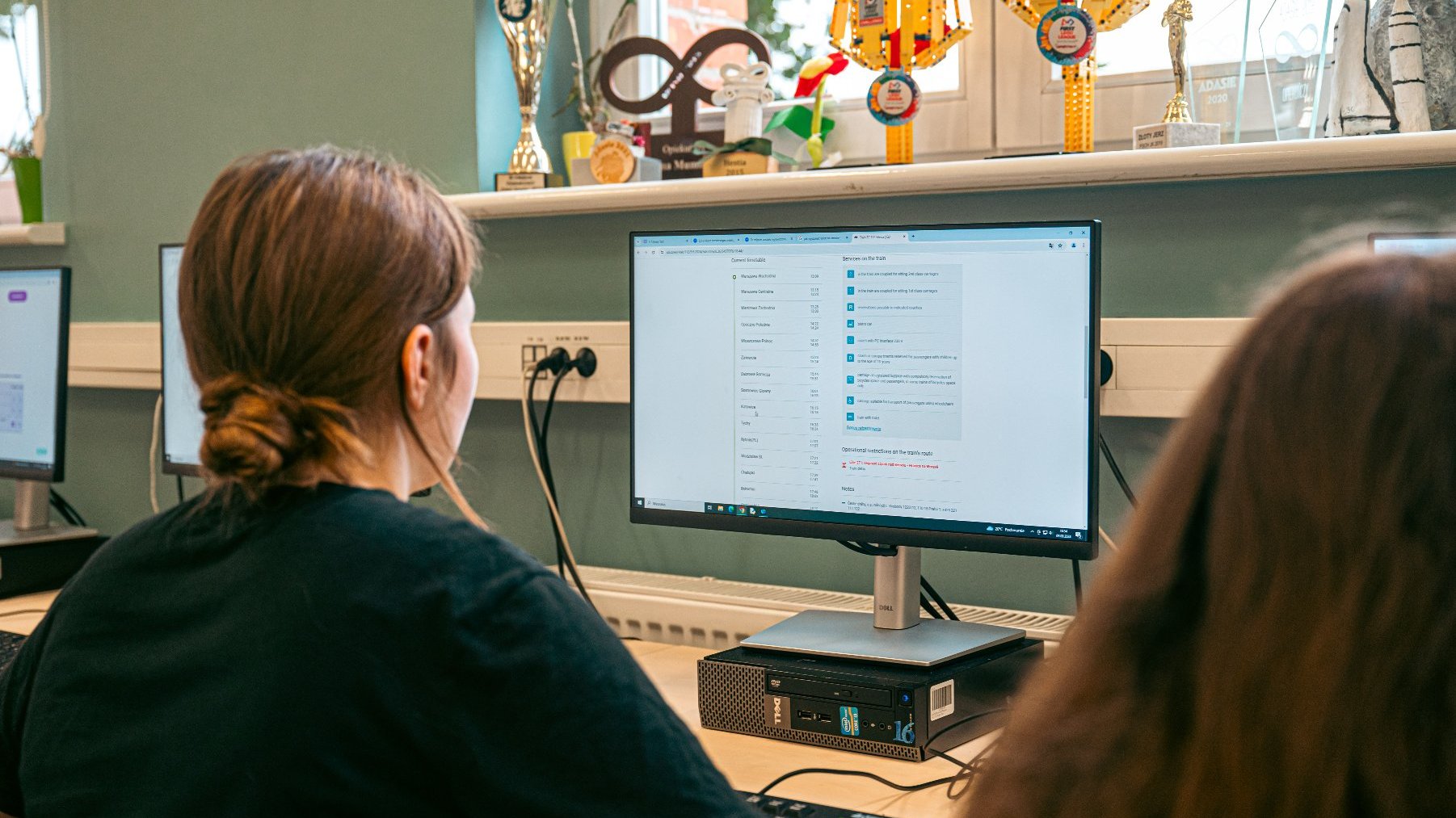 Dziewczyna siedzi tyłem do kamery przed monitorem komputerowym, pracując. Na ekranie widać otwartą aplikację. Na biurku obok komputera leży klawiatura. W tle, na parapecie, stoją liczne puchary i trofea, w tym model promu kosmicznego i statuetka robota z klocków.