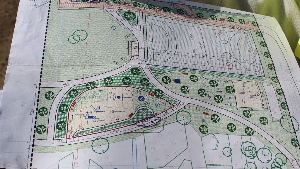 Rzut z góry przedstawiający fragment architektonicznego planu zagospodarowania terenu. Na projekcie widoczny jest plac zabaw, boisko sportowe oraz ścieżki i tereny zielone z symbolami drzew. Plan jest wydrukowany na kartce, którą trzymają dwie dłonie.
