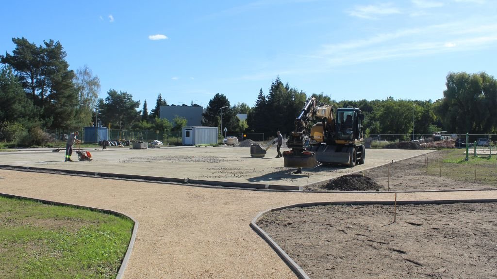 Teren rekreacyjny w trakcie budowy lub przebudowy. Na pierwszym planie widoczna żwirowa ścieżka z obrzeżami, rozwidlająca się w kształt litery "Y". W głębi, na utwardzonym placu, stoi żółta koparka oraz dwóch pracowników. W tle drzewa i budynki. W prawym dolnym rogu widoczny symbol ZZM.