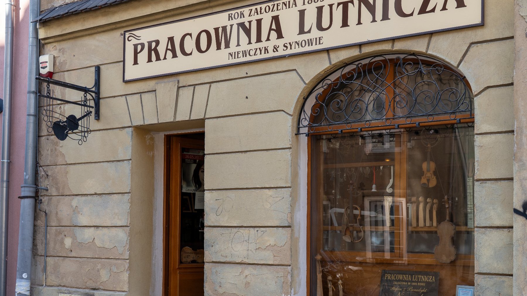 Wejście do Pracowni Lutniczej w zabytkowym budynku o beżowej elewacji. Nad wejściem widoczny duży szyld z napisem: "ROK ZAŁOŻENIA 1885 PRACOWNIA LUTNICZA NIEWCZYK & SYNOWIE". Po prawej stronie okno wystawowe z łukowym nadprożem i kratą, za którym widać skrzypce i instrumenty muzyczne. W lewym górnym rogu na ścianie wisi metalowy kuty szyld w kształcie skrzypiec.