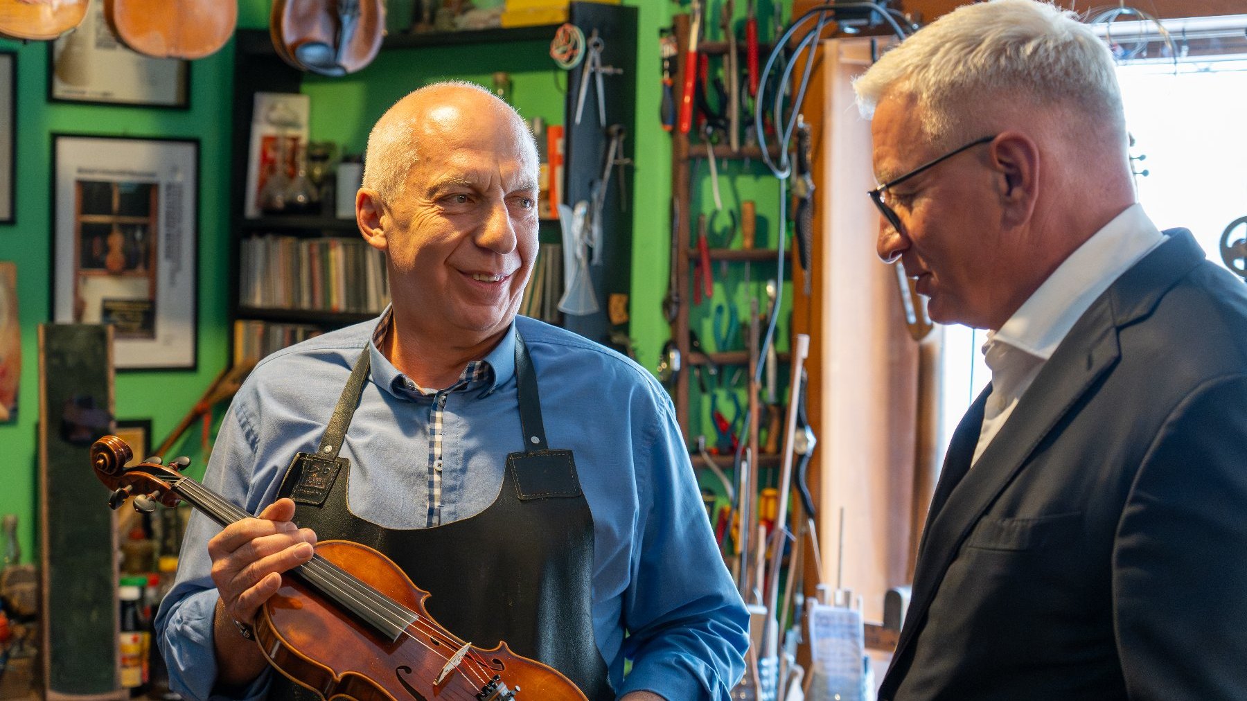 W warsztacie lutniczym dwóch mężczyzn rozmawia. Mężczyzna po lewej, ubrany w niebieską koszulę i czarny fartuch, uśmiecha się i trzyma w rękach skrzypce. Mężczyzna po prawej jest w ciemnym garniturze i okularach. W tle widać zieloną ścianę i liczne narzędzia lutnicze.